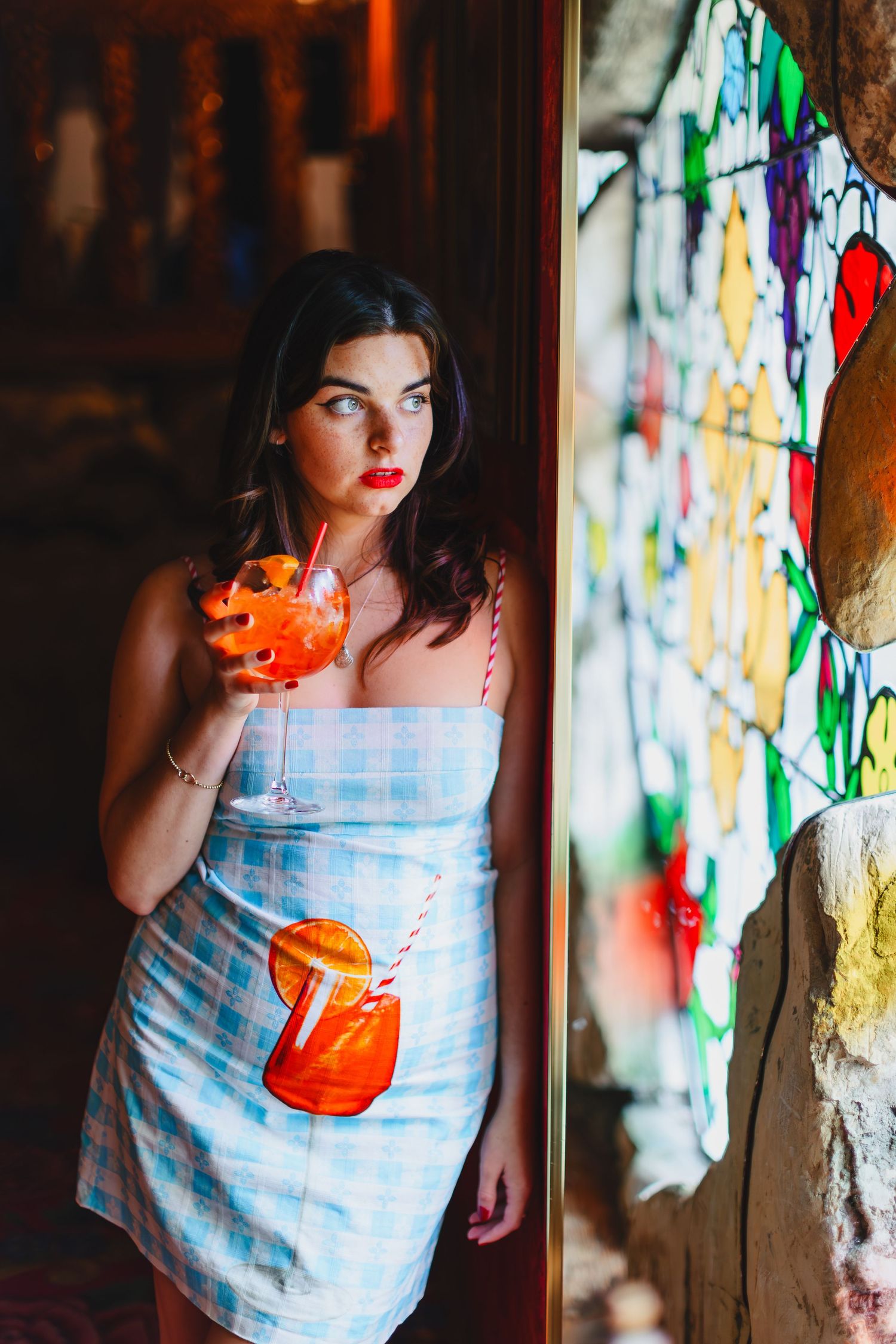 Woman in blue dress holds orange cocktail near stained glass window.