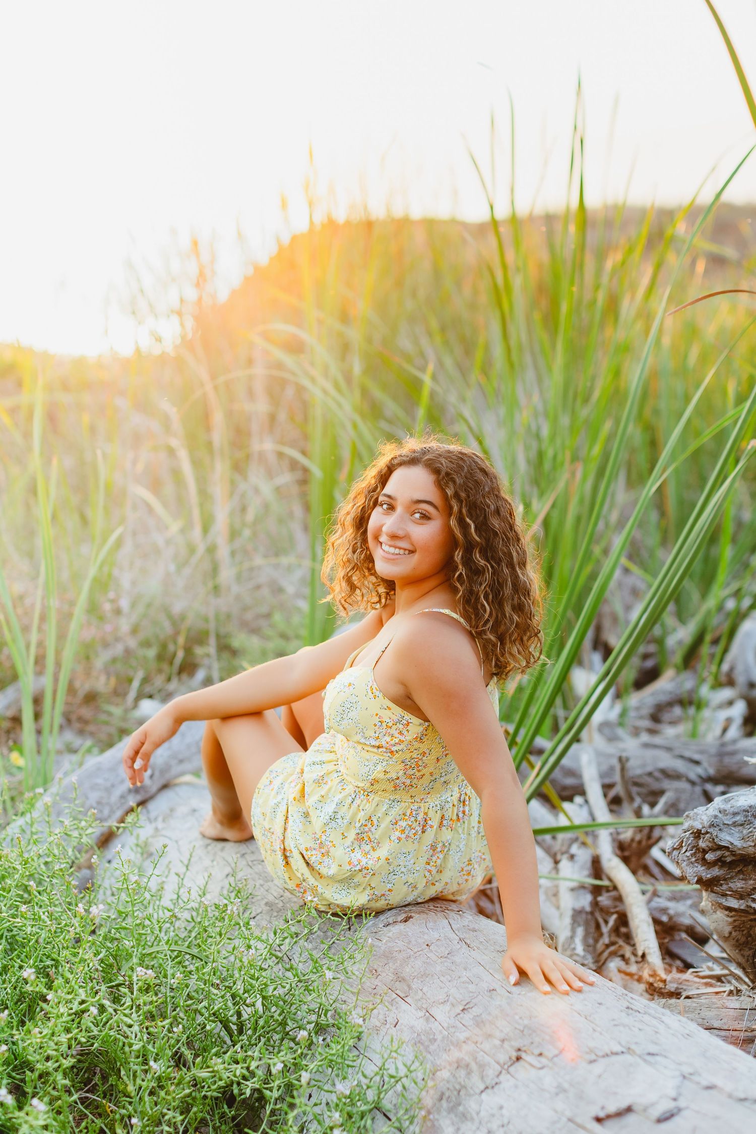 Young woman with curly hair in a yellow floral sundress sitting on rocks surrounded by tall green grass in golden sunlight.