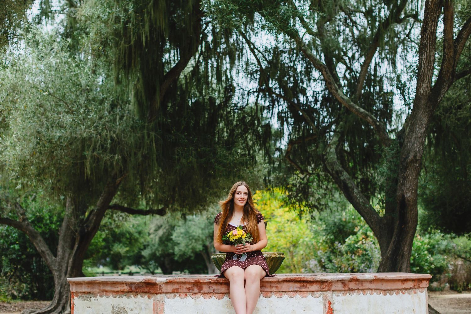 A person sits on a brick wall surrounded by lush trees and greenery in a serene outdoor setting.