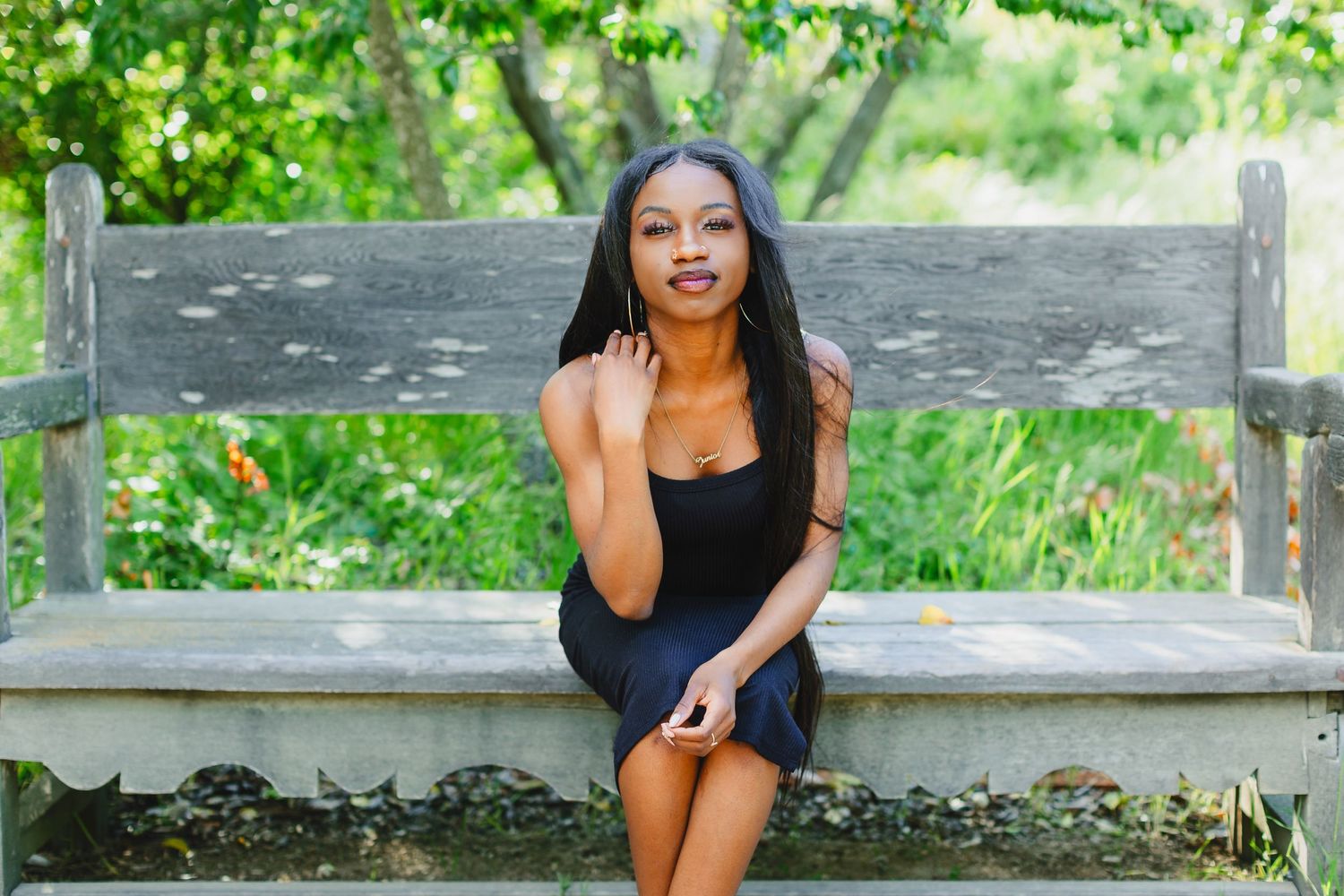 Woman in black dress sits casually on wooden bench in sunny garden setting.