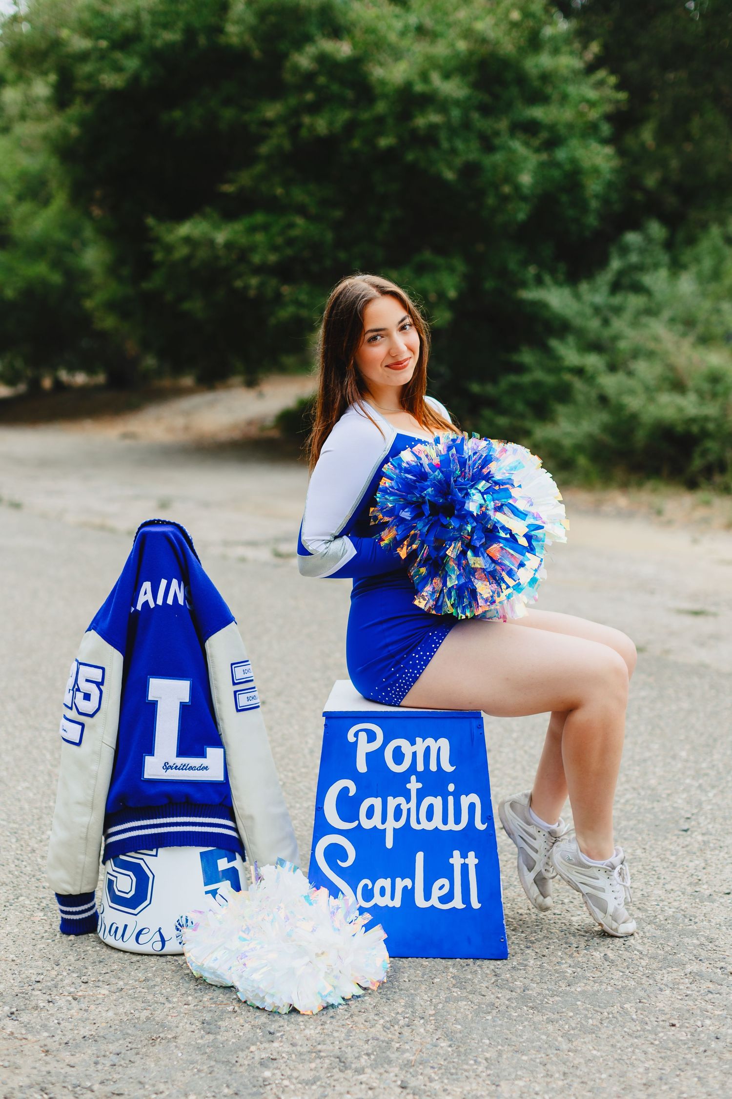High school pom captain Scarlett poses in blue cheerleading uniform with pom-poms and a letter jacket outdoors.