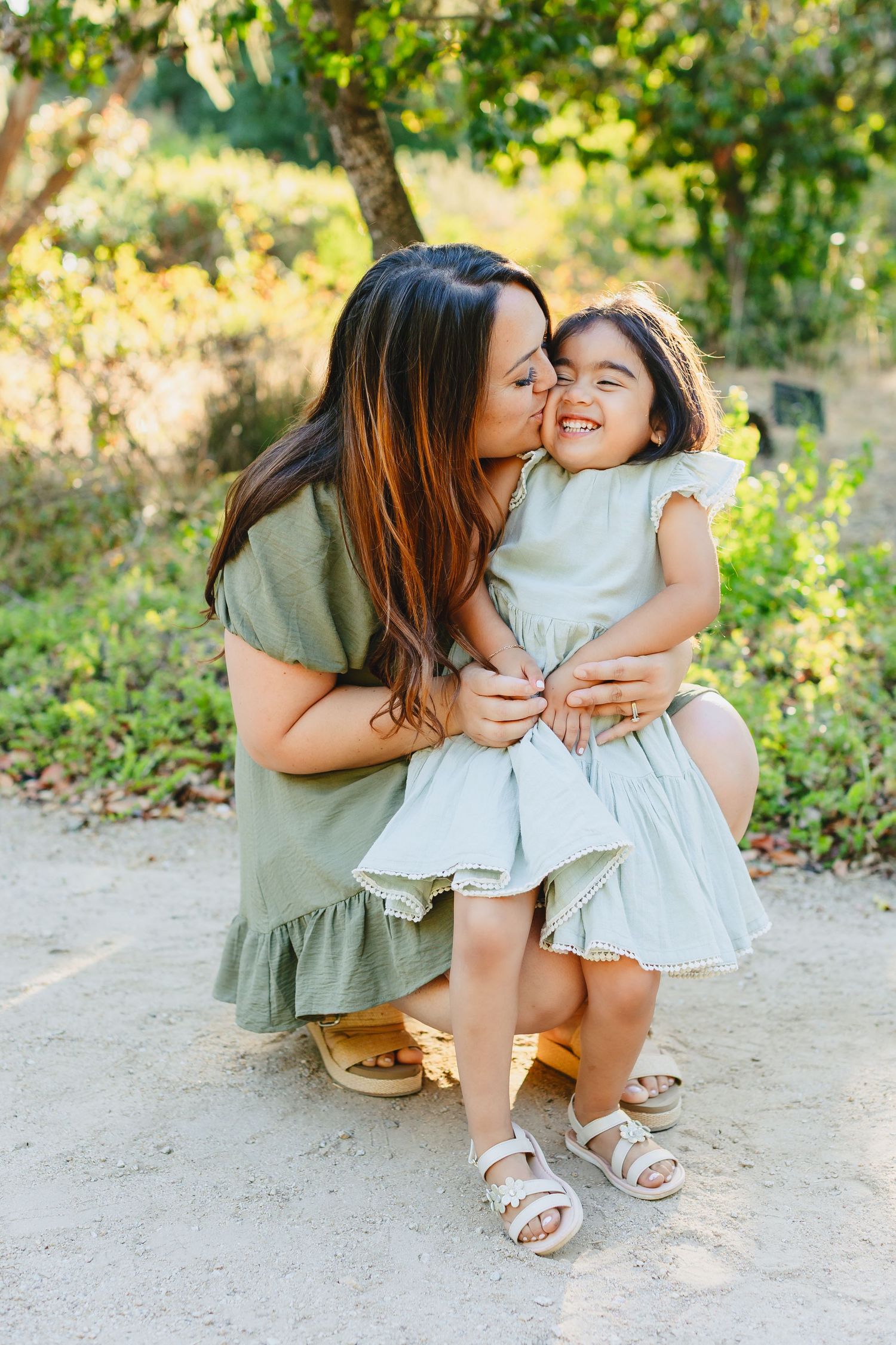 A tender family moment captured in nature with soft sunlight filtering through green foliage in the background.
