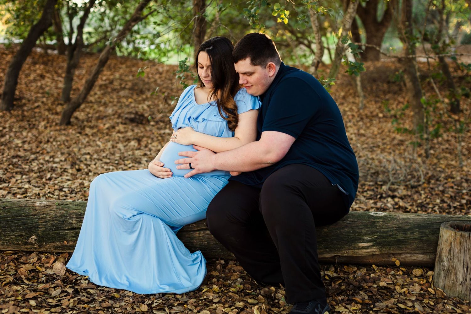 A romantic couple in matching blue outfits sits together on a fallen tree trunk in a wooded setting during autumn.