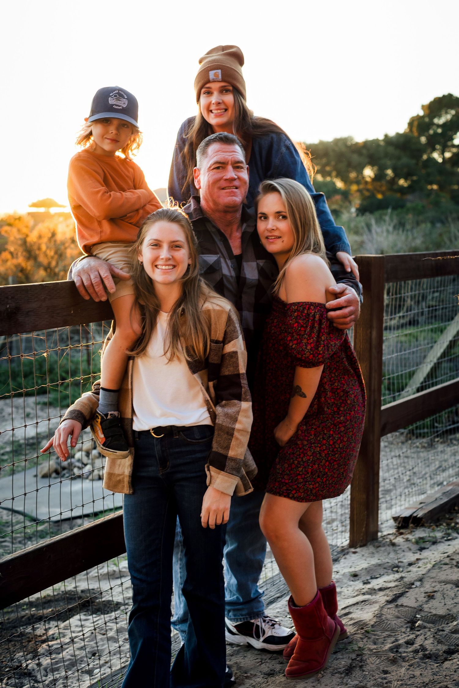 A family poses together at sunset by a wooden fence in casual fall attire on a rustic farm path.