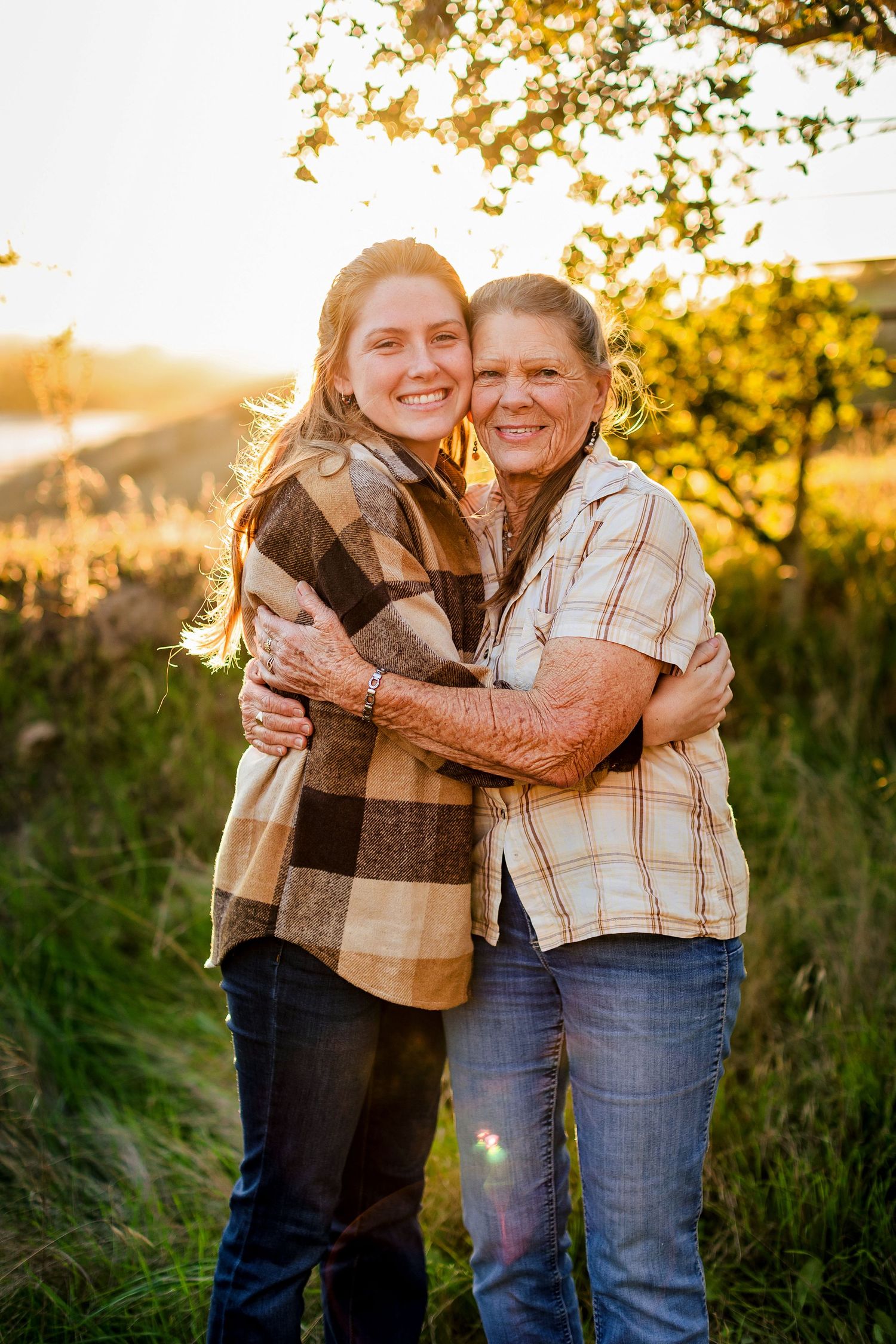 Two people embrace warmly outdoors during golden hour in a field with tall grass and golden sunlight.