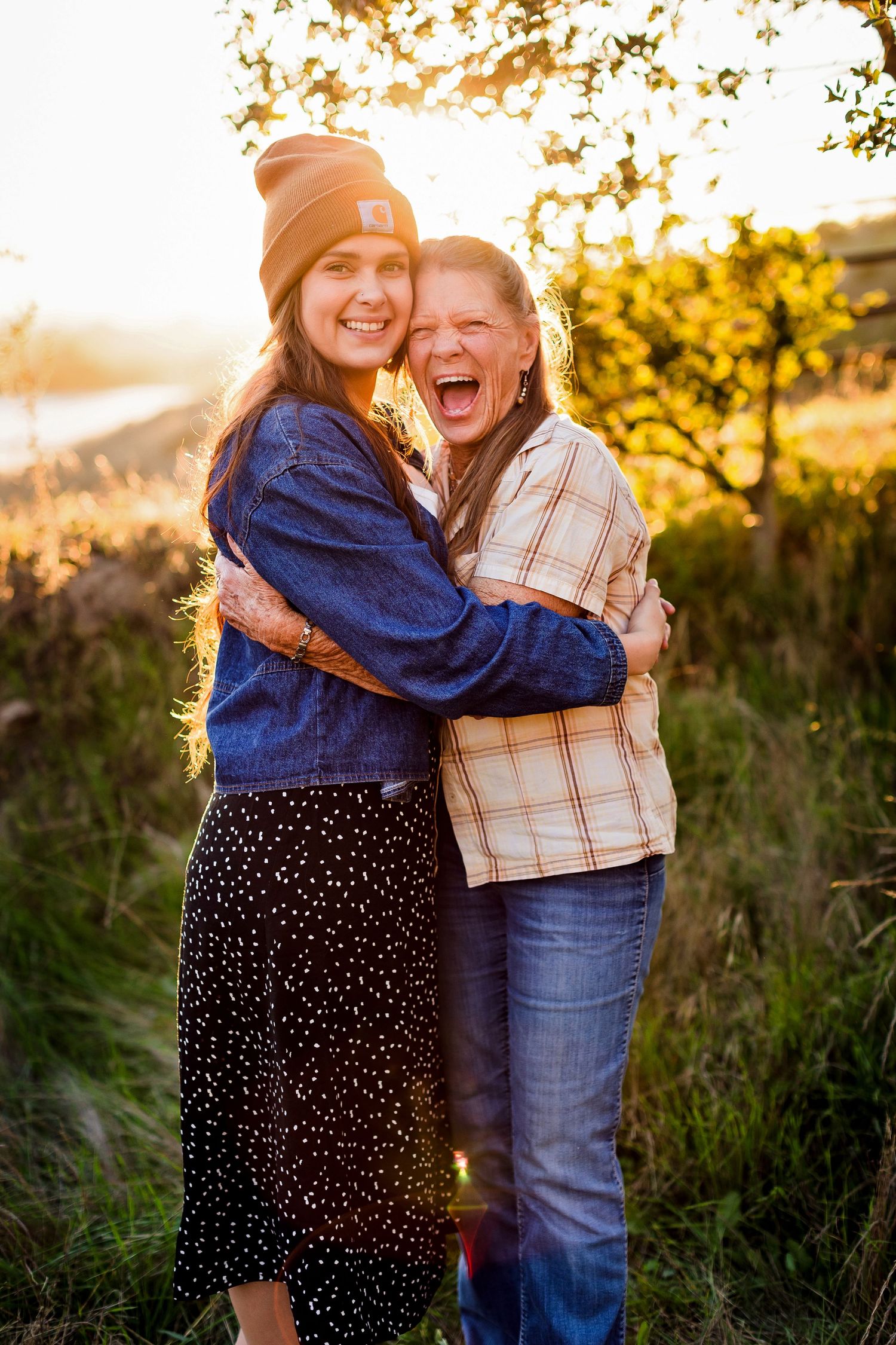Two people embrace and laugh together in a golden sunset field with tall grass and warm backlighting.