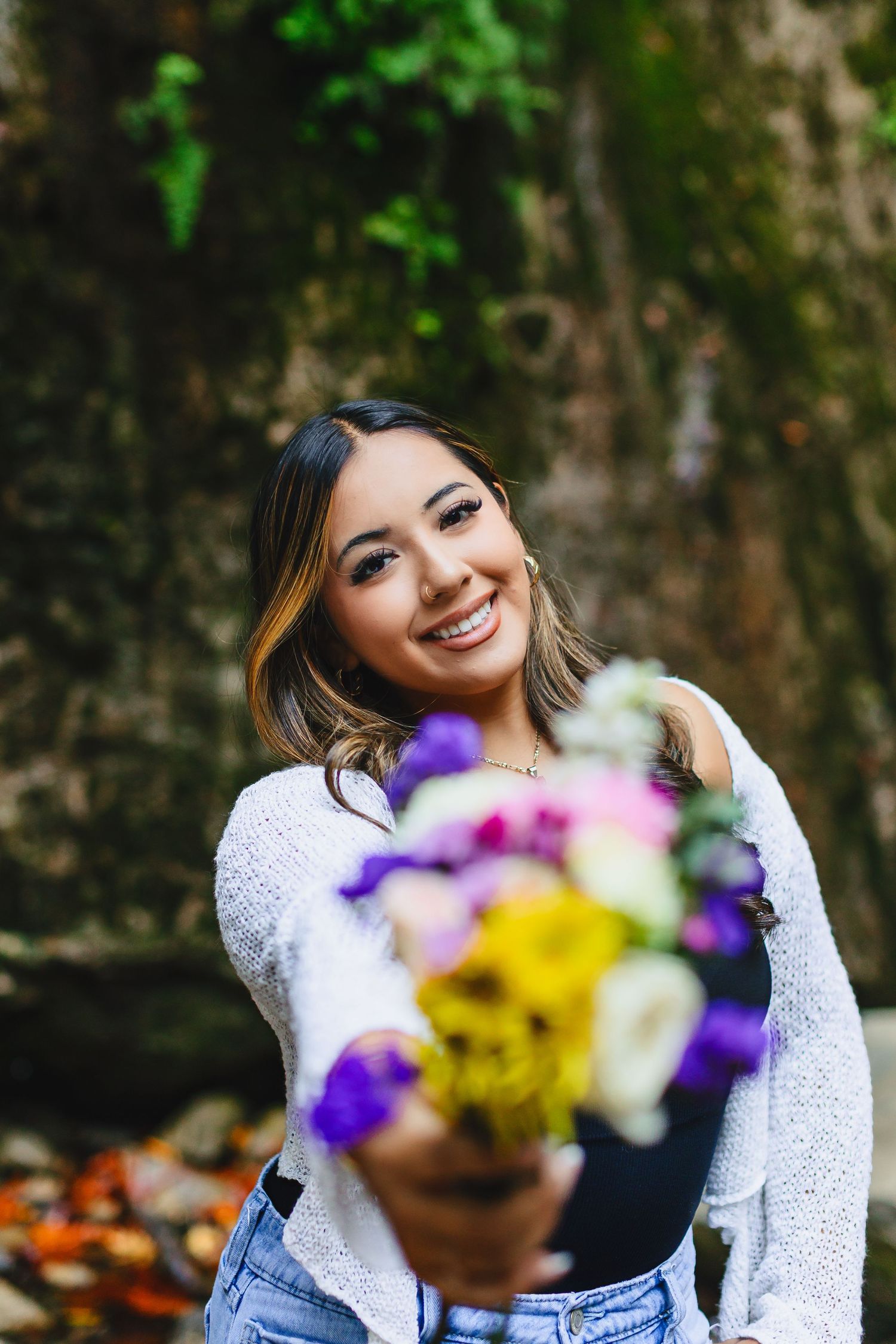 Smiling young woman in white sweater holding a colorful bouquet of flowers outdoors near a mossy stone wall.
