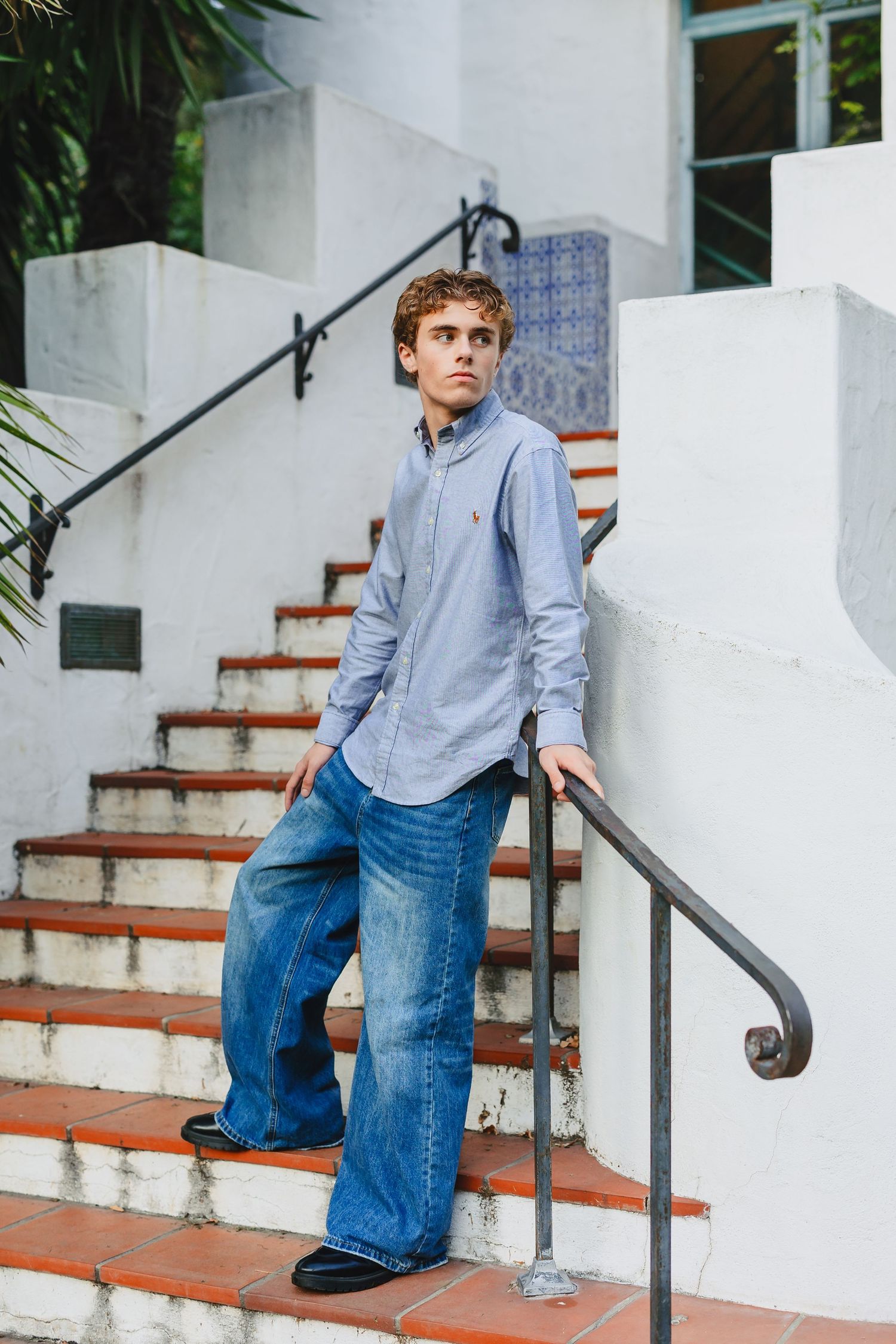 Young man in wide-leg jeans and blue Oxford shirt poses on brick stairs with white stucco building backdrop.
