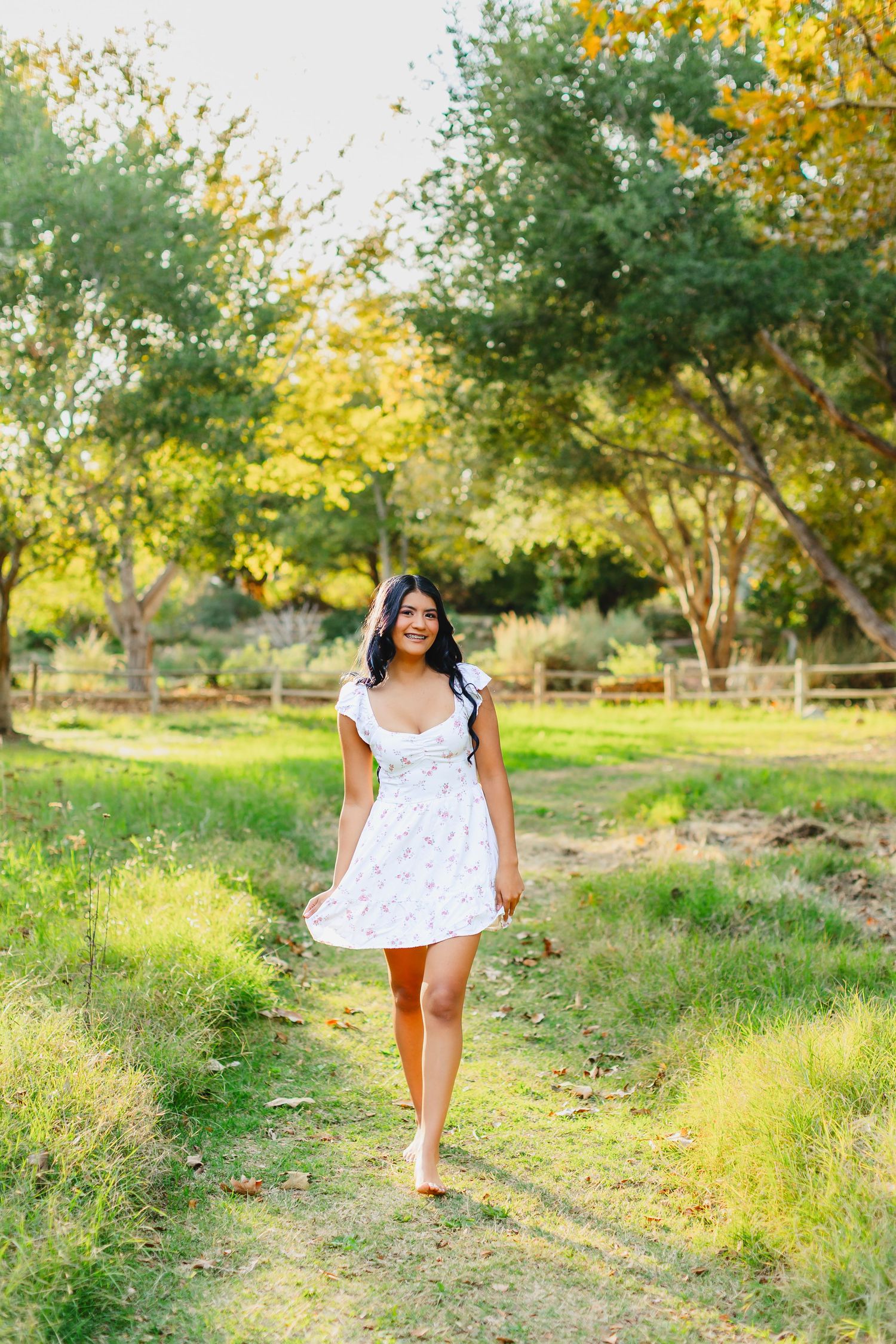 Young woman in white floral dress walking barefoot on a grass path in a sunny green park.