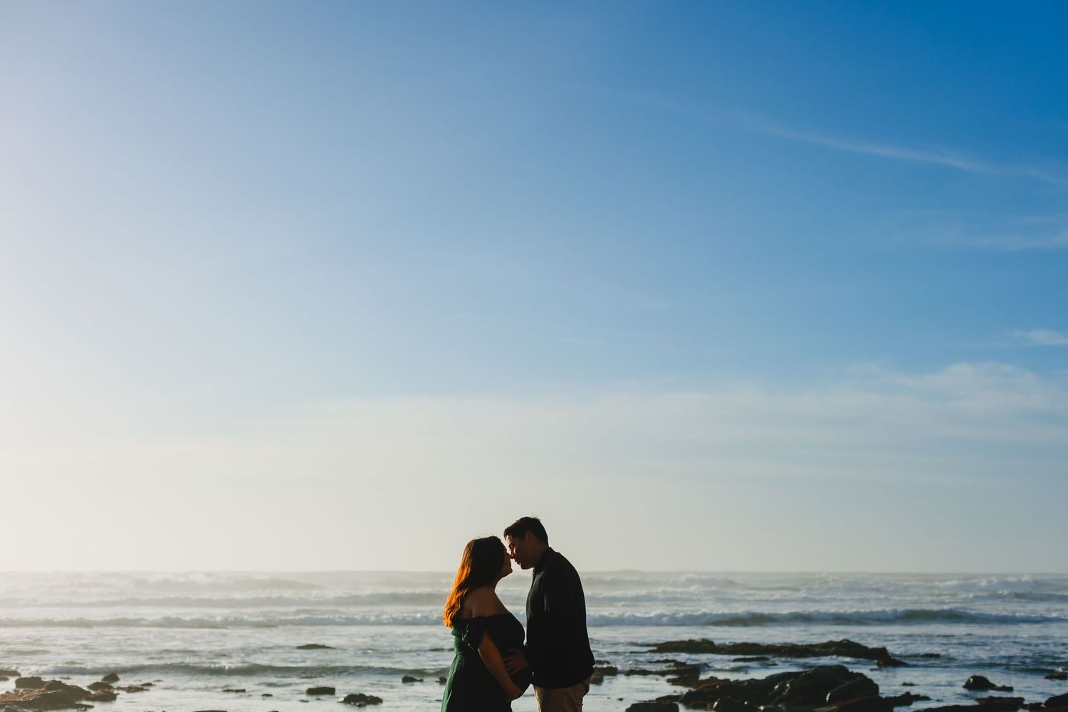 Two silhouetted figures stand close together on rocky beach against dramatic bright ocean horizon and blue sky.