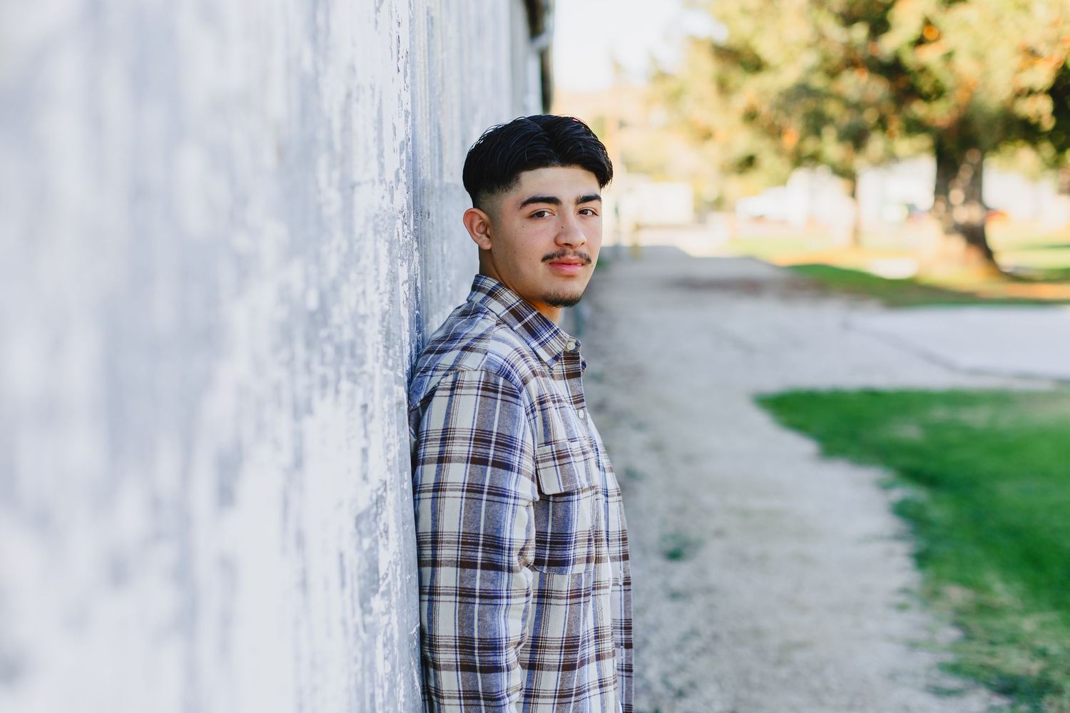 Person in plaid shirt and black cap leans against white wall on sunny path next to grassy area.