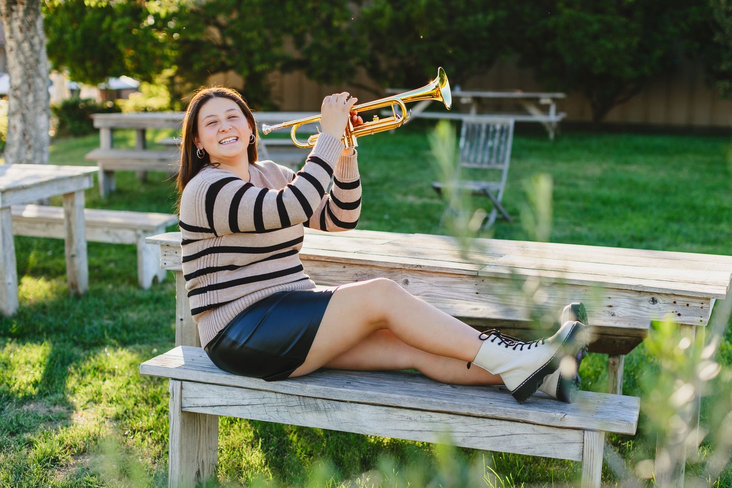 Smiling young woman sitting on wooden bench holding a gold trumpet in an outdoor park setting.