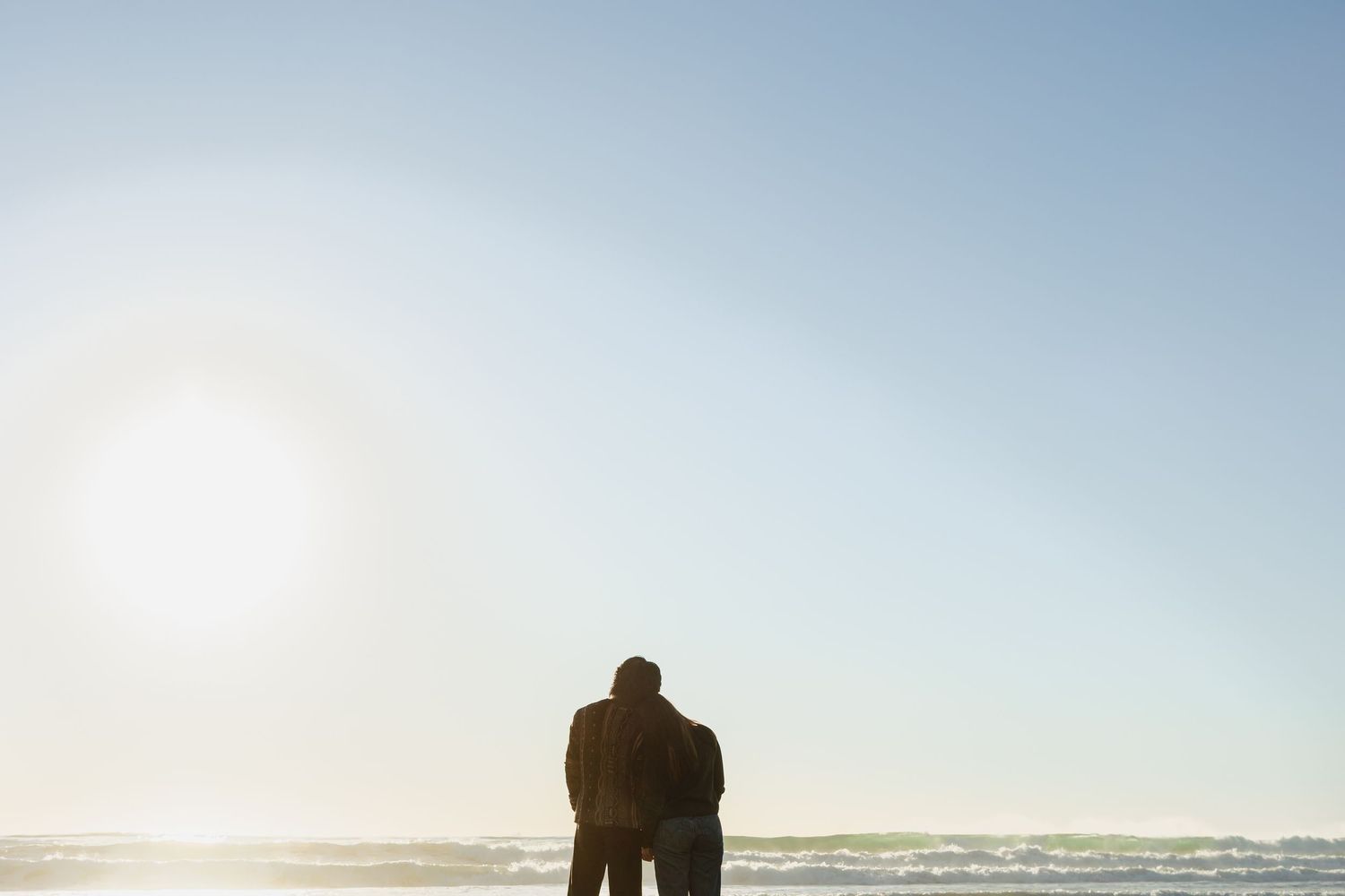 Couple standing together on a beach at sunset, silhouetted against a bright sky and calm ocean waves.