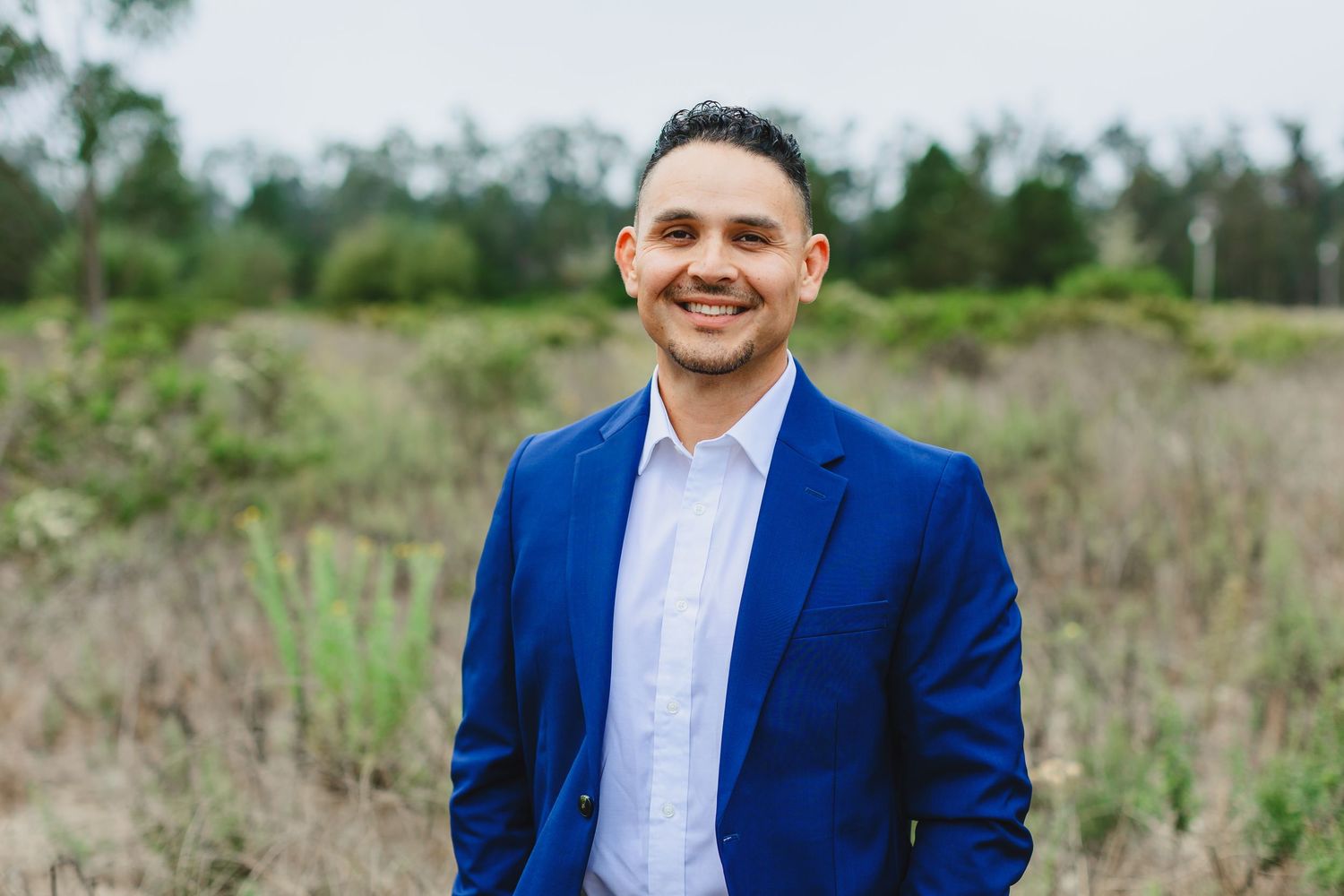 A person in a bright blue suit and white dress shirt stands outdoors in a grassy field on a sunny day.
