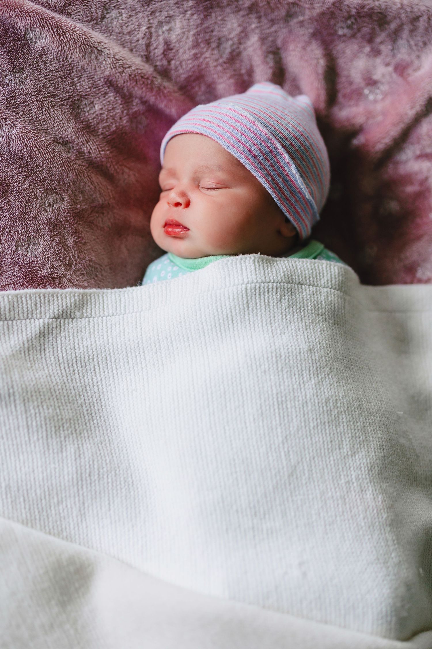 Peaceful newborn baby sleeping soundly, wrapped in a white swaddle blanket and wearing a striped hospital hat.
