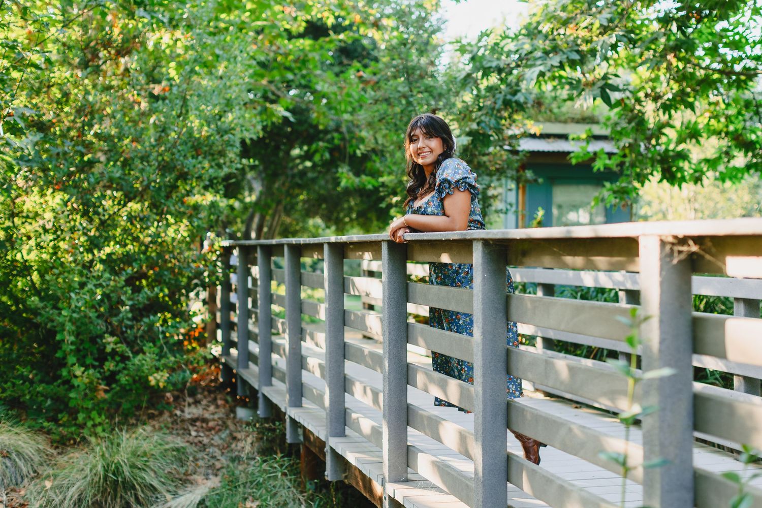 Someone in a blue shirt and sunglasses leans against wooden railing of outdoor bridge surrounded by green foliage.