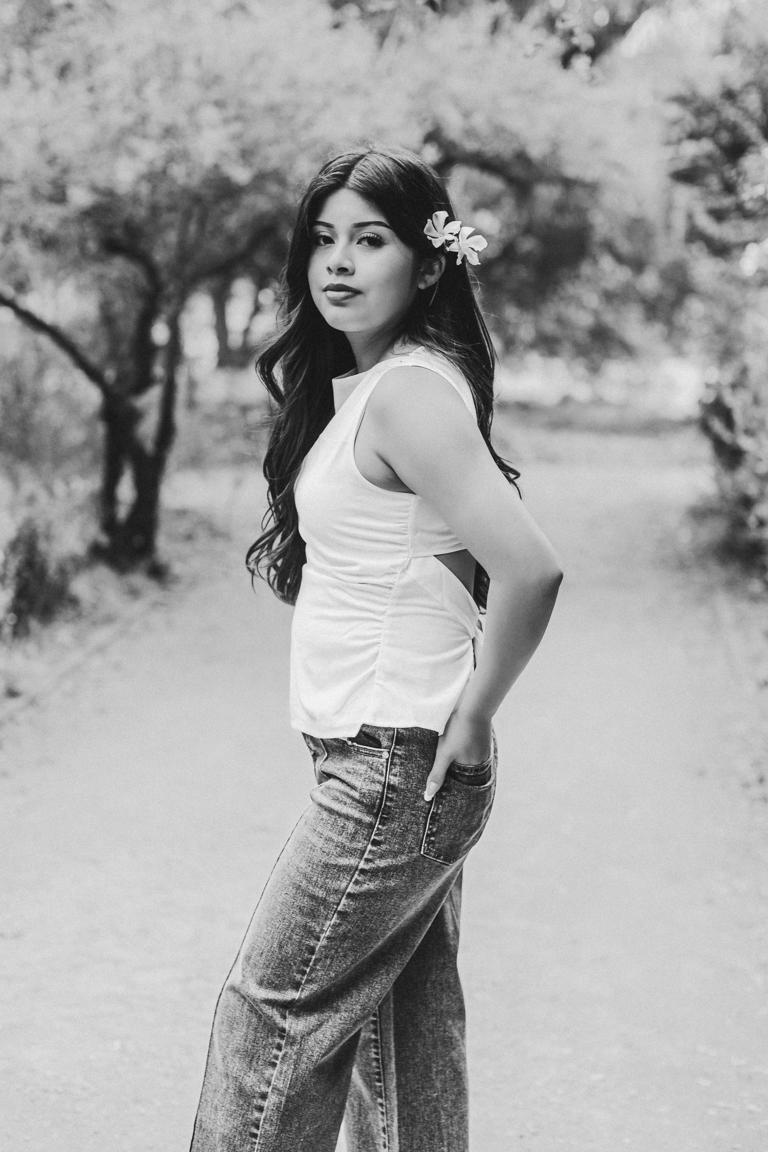 Black and white photo of a young woman with a flower in her hair, wearing a white top and jeans outdoors.