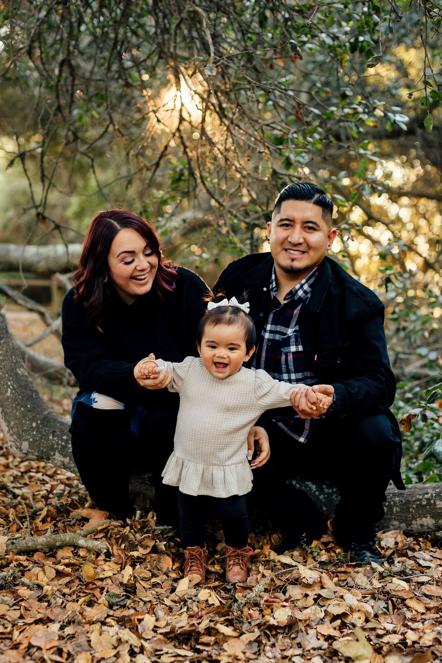 Family of three sits together on fallen autumn leaves while sharing joyful moments in an outdoor setting.