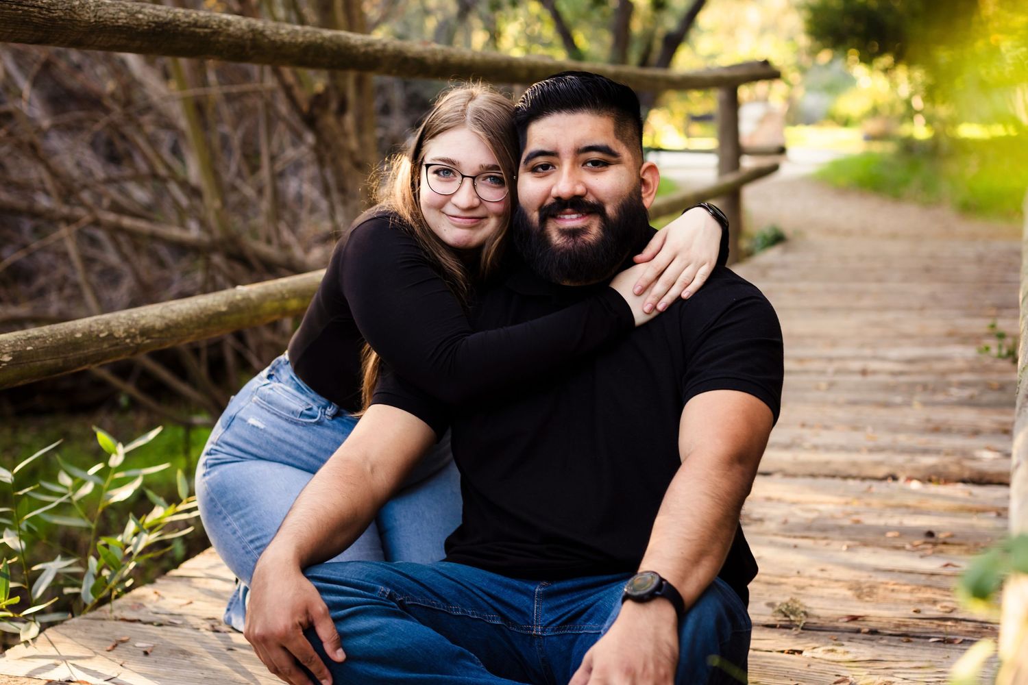Two people in blue jeans and black shirts pose together on wooden stairs surrounded by greenery.