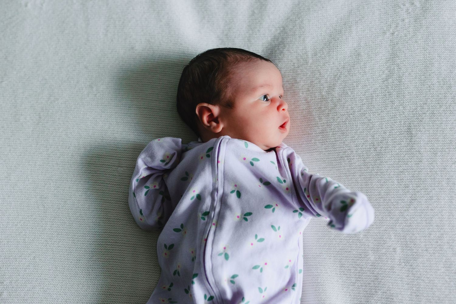 Newborn baby lying on back in white floral onesie on light blue blanket, looking to the side.