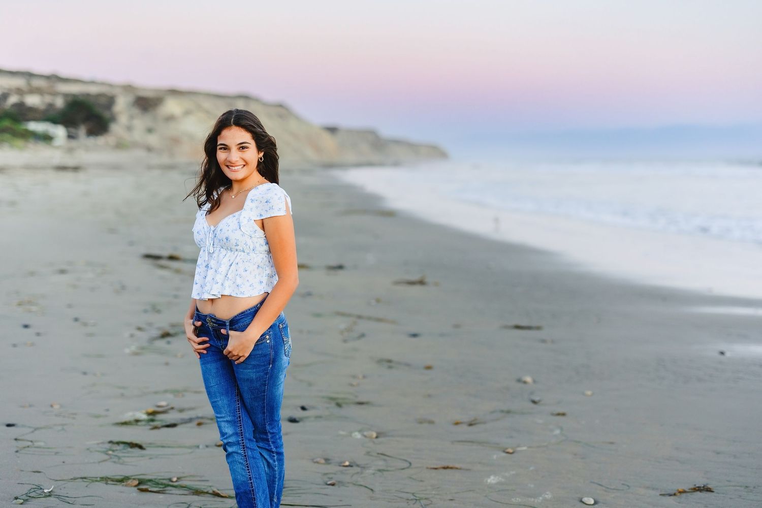 Young woman smiling on a sandy beach at sunset, wearing jeans and a white top, holding a camera.