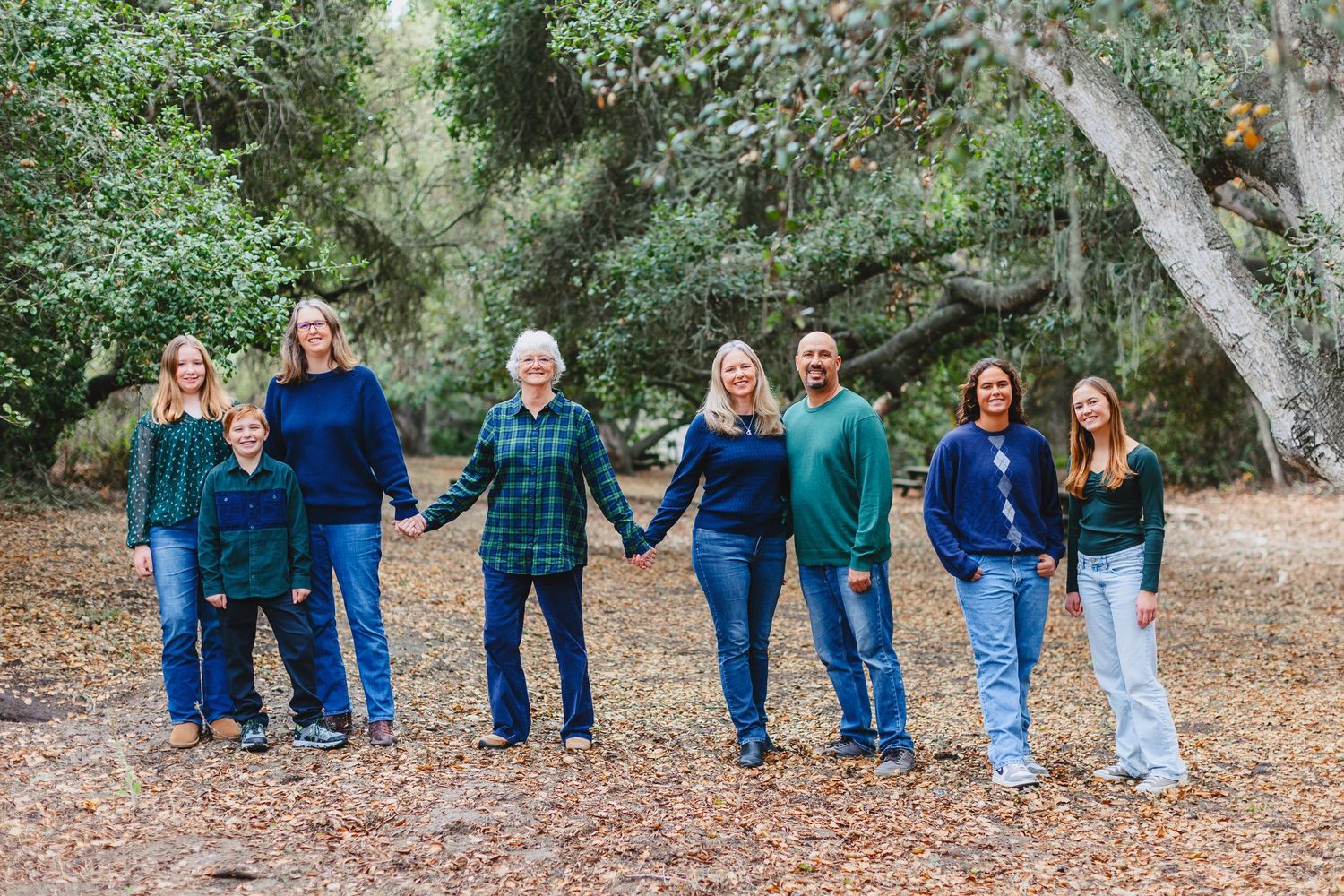 A group in coordinating blue and teal outfits holds hands while standing on fallen leaves in a wooded outdoor setting.
