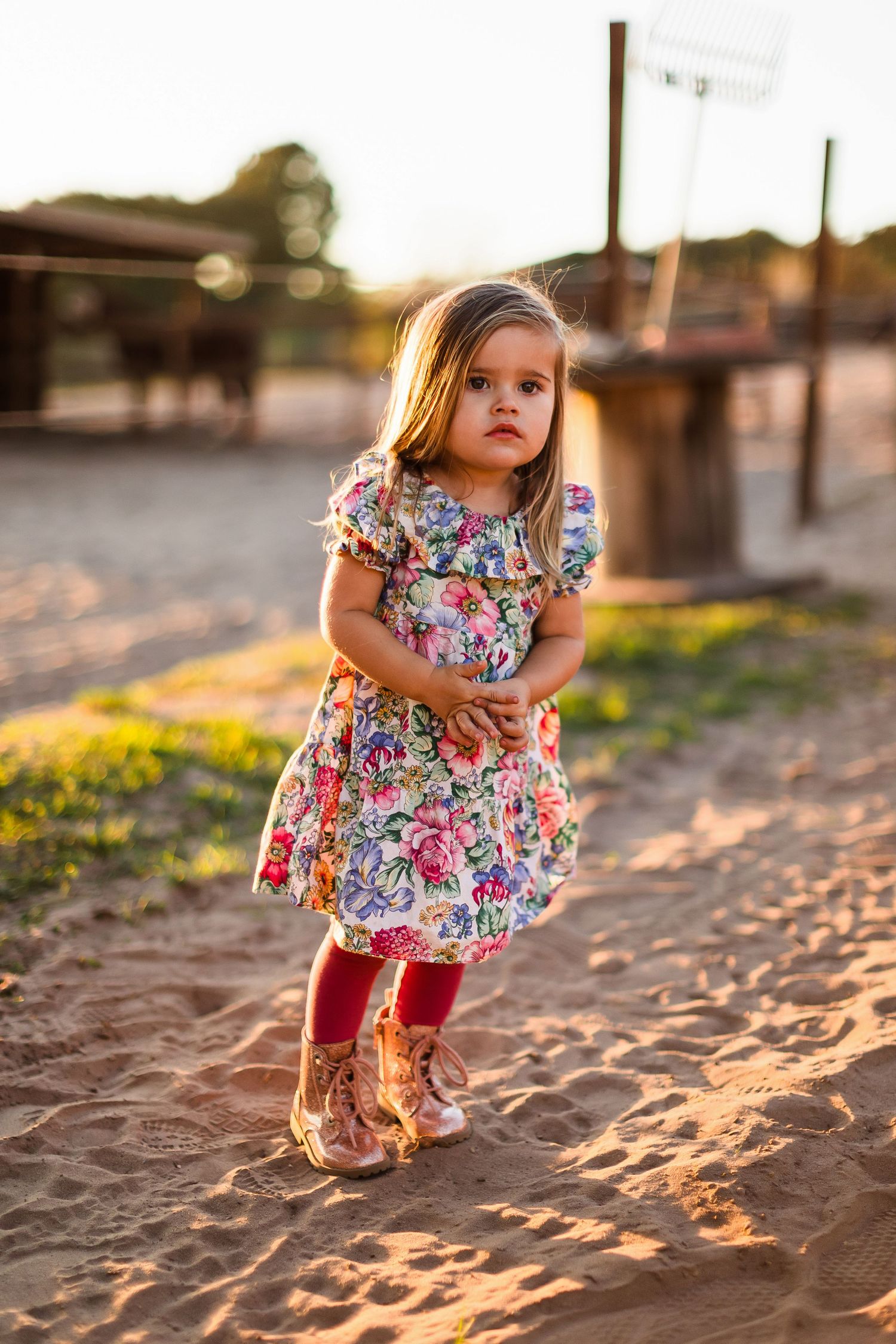 A small child in a floral dress and red leggings plays joyfully at sunset in a sandy outdoor area.