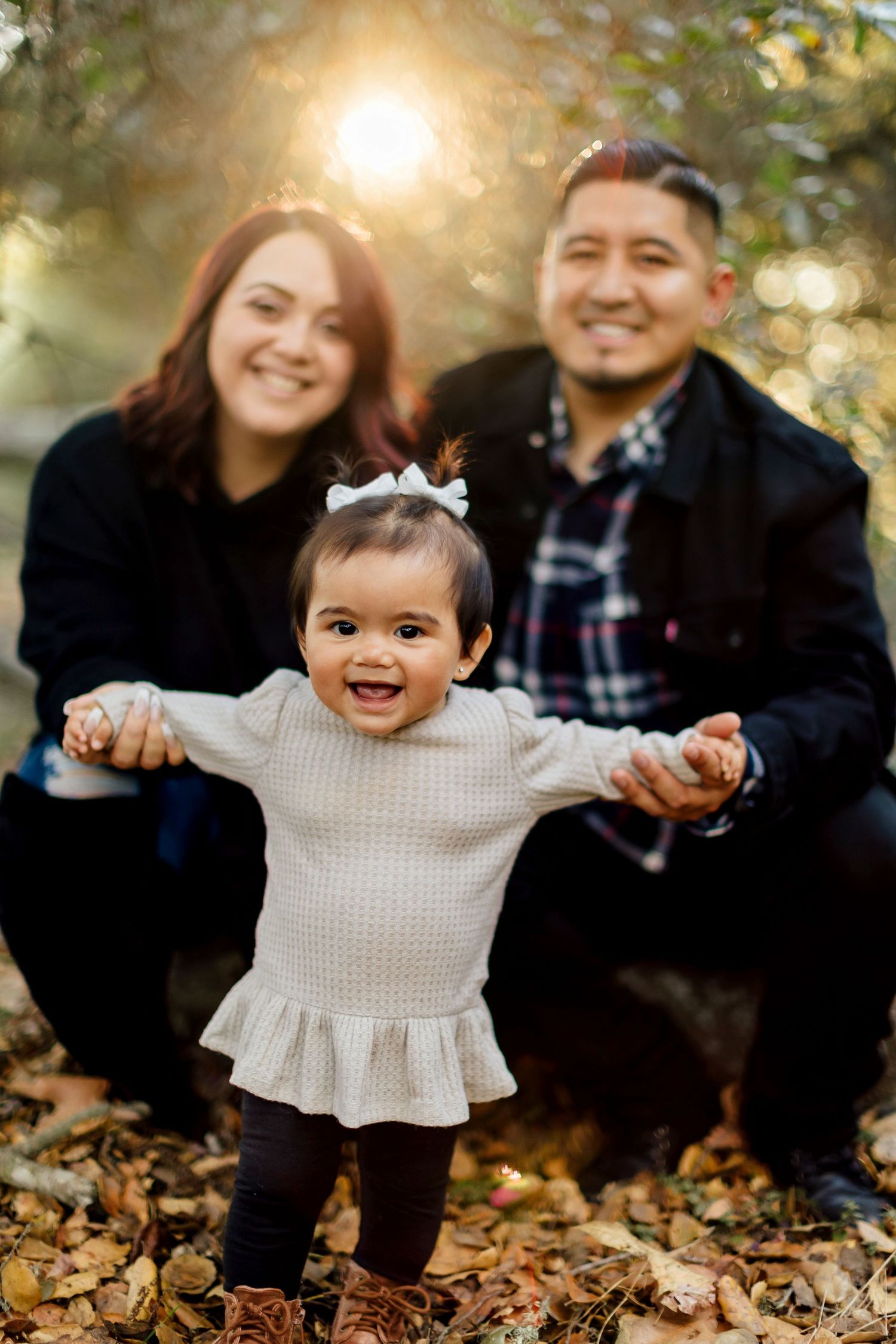 A family sits together on autumn leaves while a happy toddler in a white dress plays during a fall photoshoot.