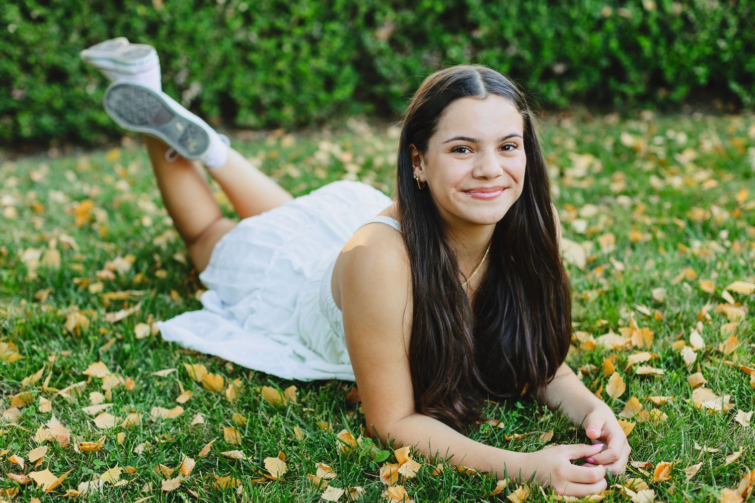 Teen girl in white dress lying on grass with autumn leaves for senior portrait photo.