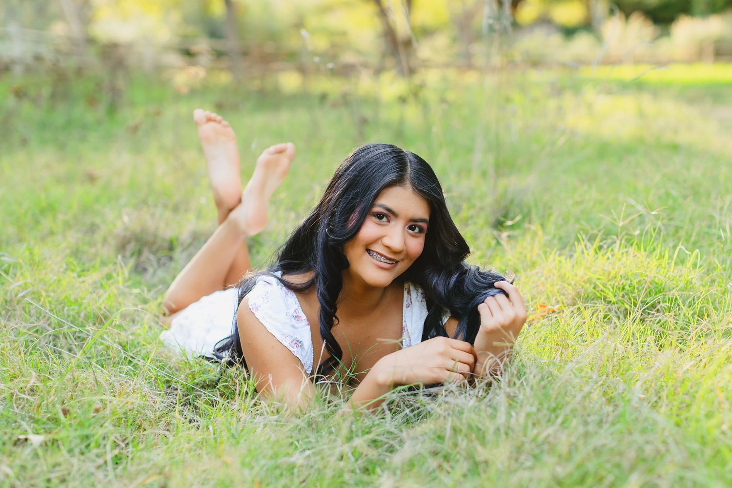 A young woman in white clothing lies in a grassy meadow on a sunny day while taking photos with a camera.