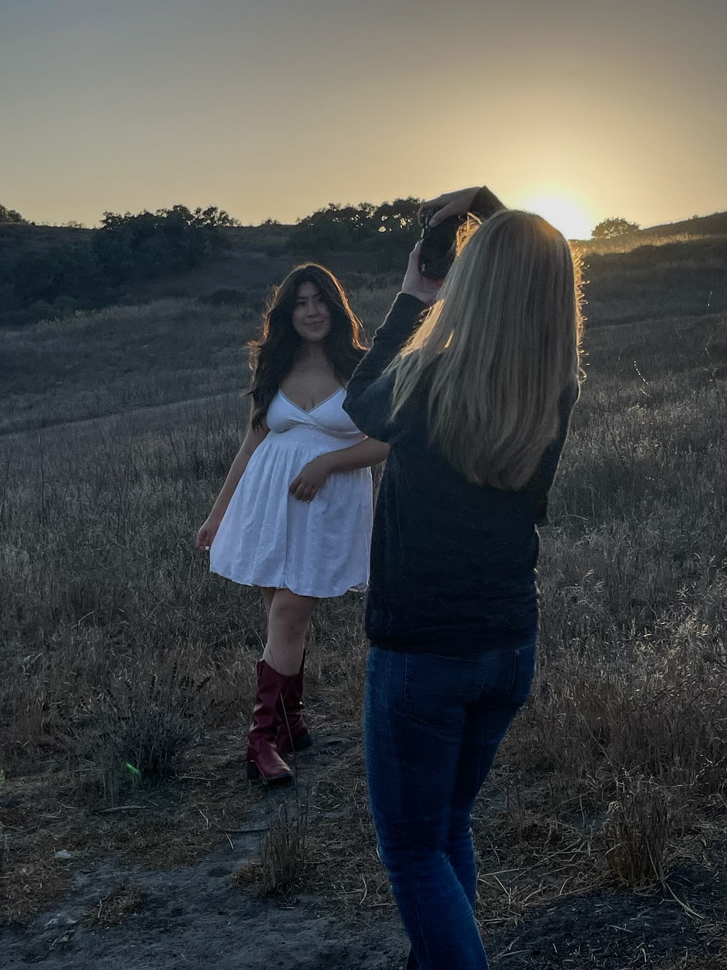 Blonde photographer shoots young woman in white dress and red boots at golden sunset.