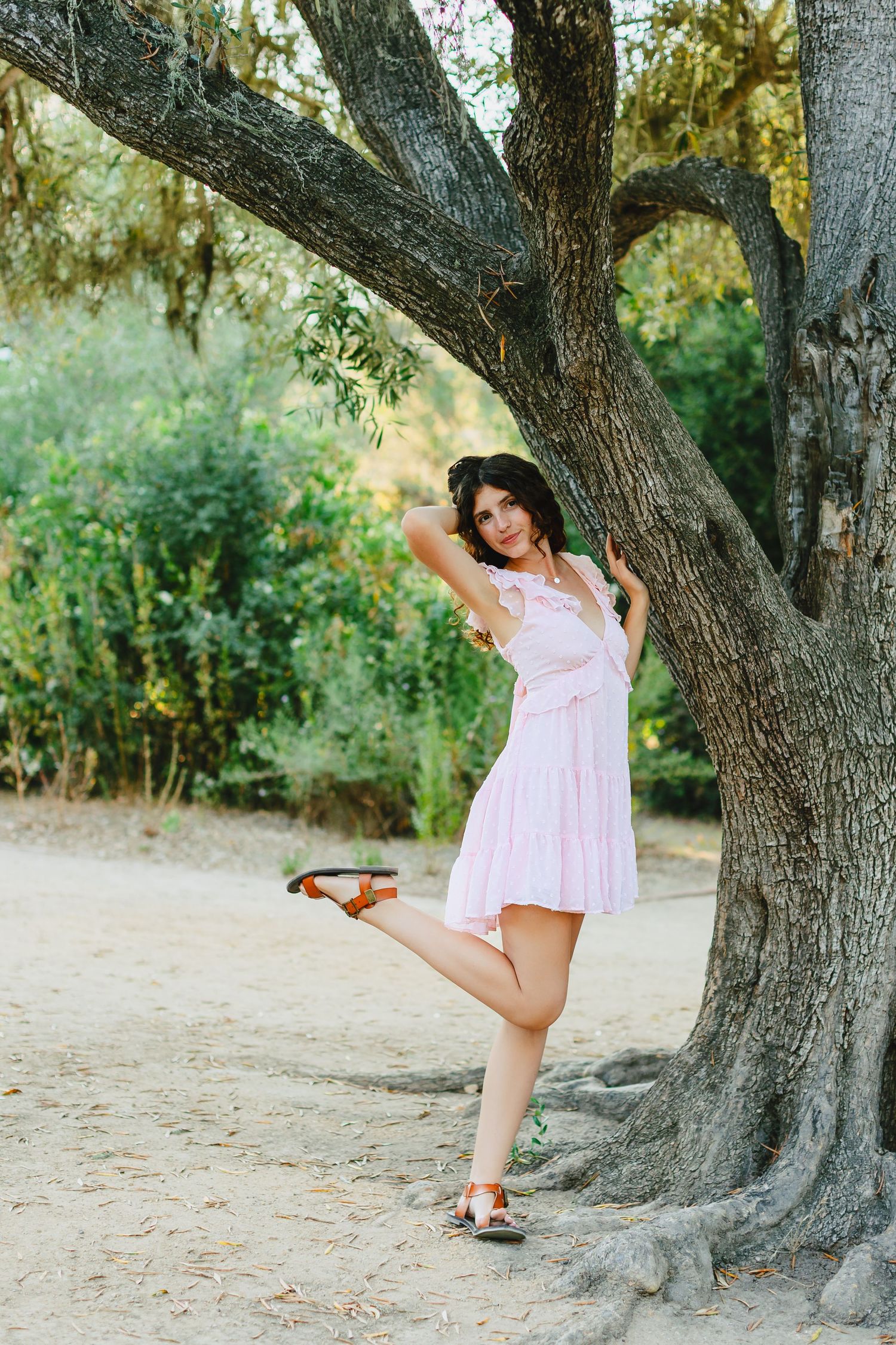 Woman in pink dress posing gracefully against a large tree trunk in a lush green outdoor setting.