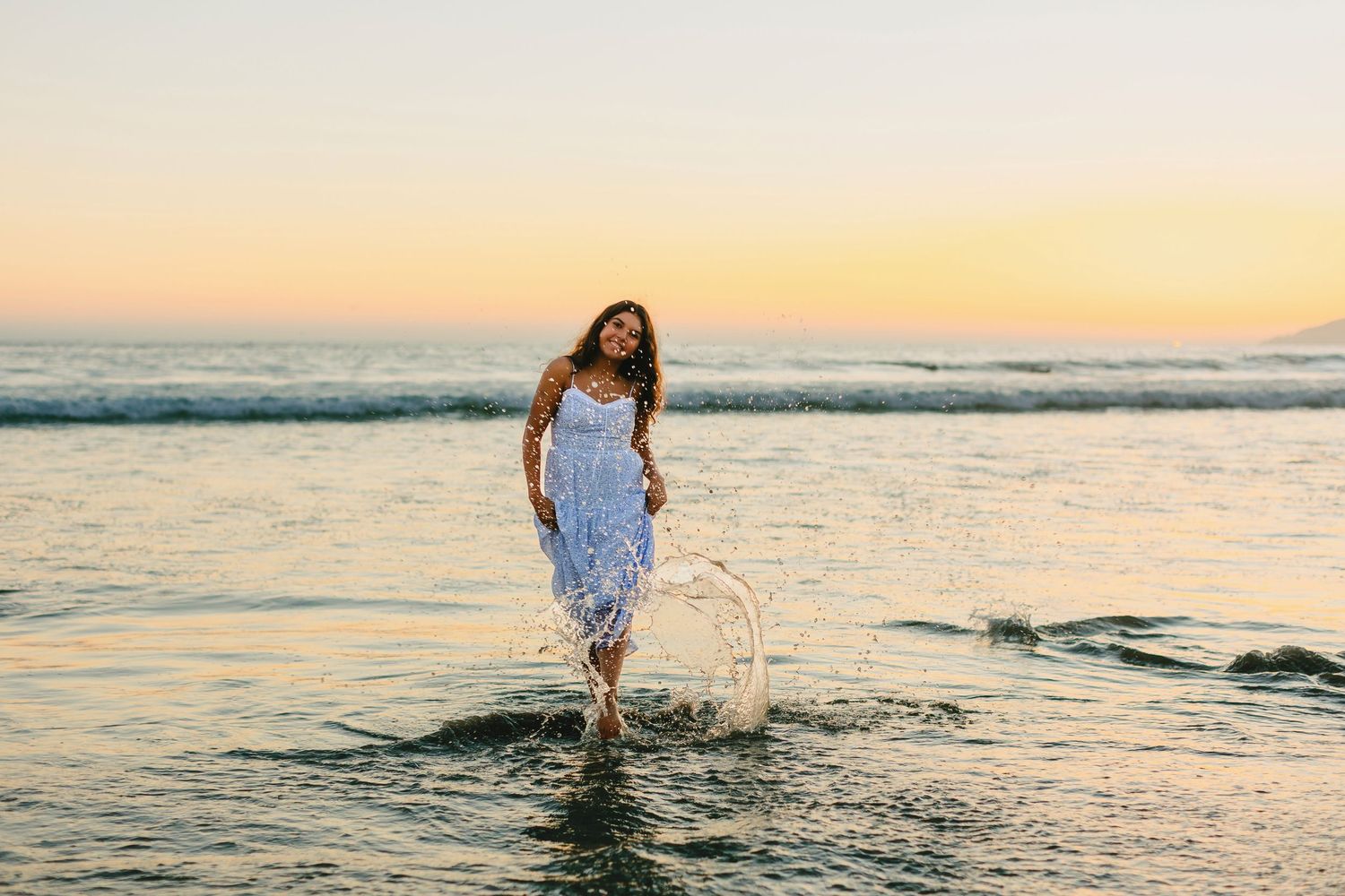 A person in a white dress splashes through shallow ocean water at sunset along a scenic beach.
