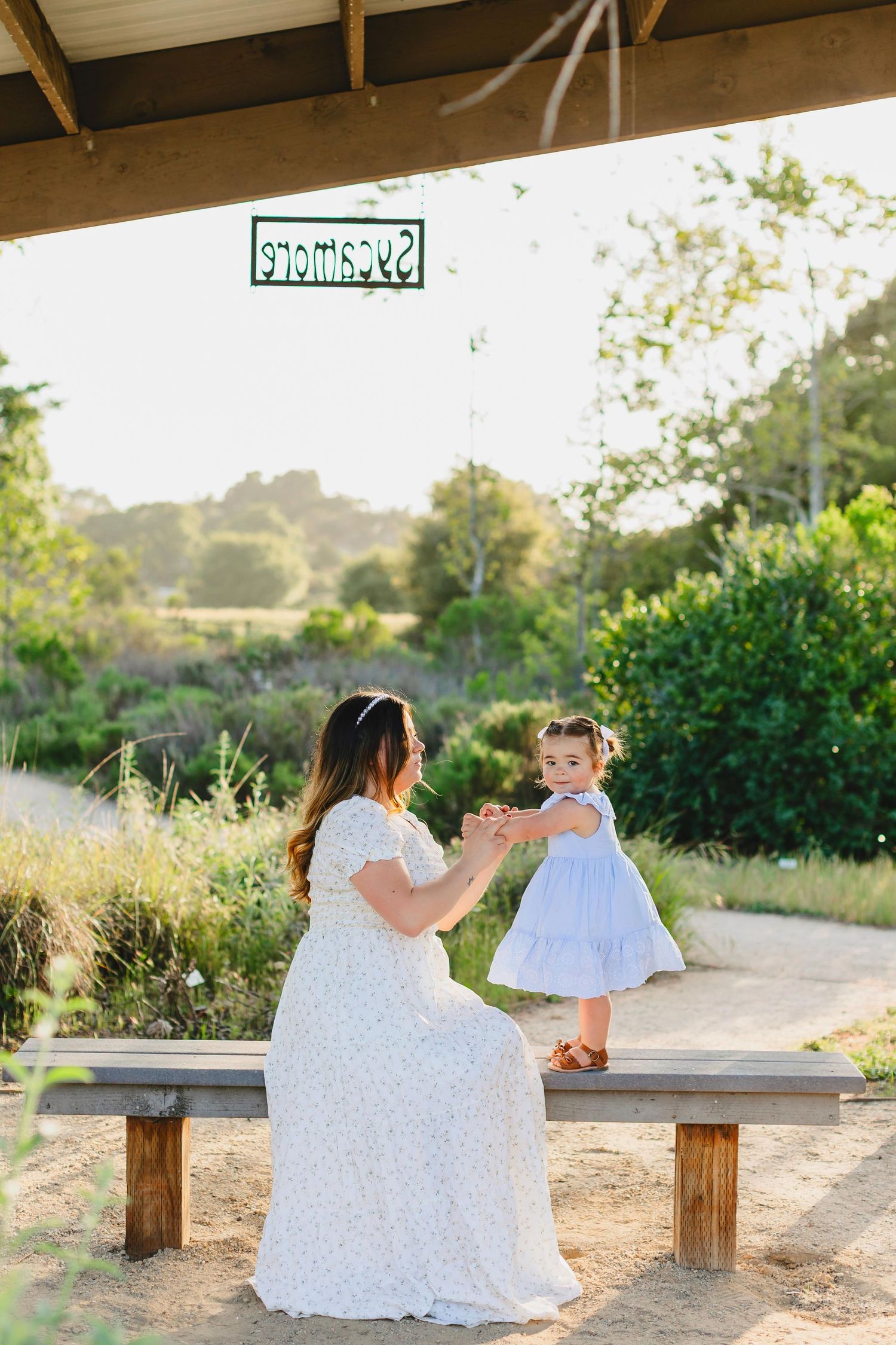 Woman and child in white dresses sitting on wooden bench in outdoor garden setting.