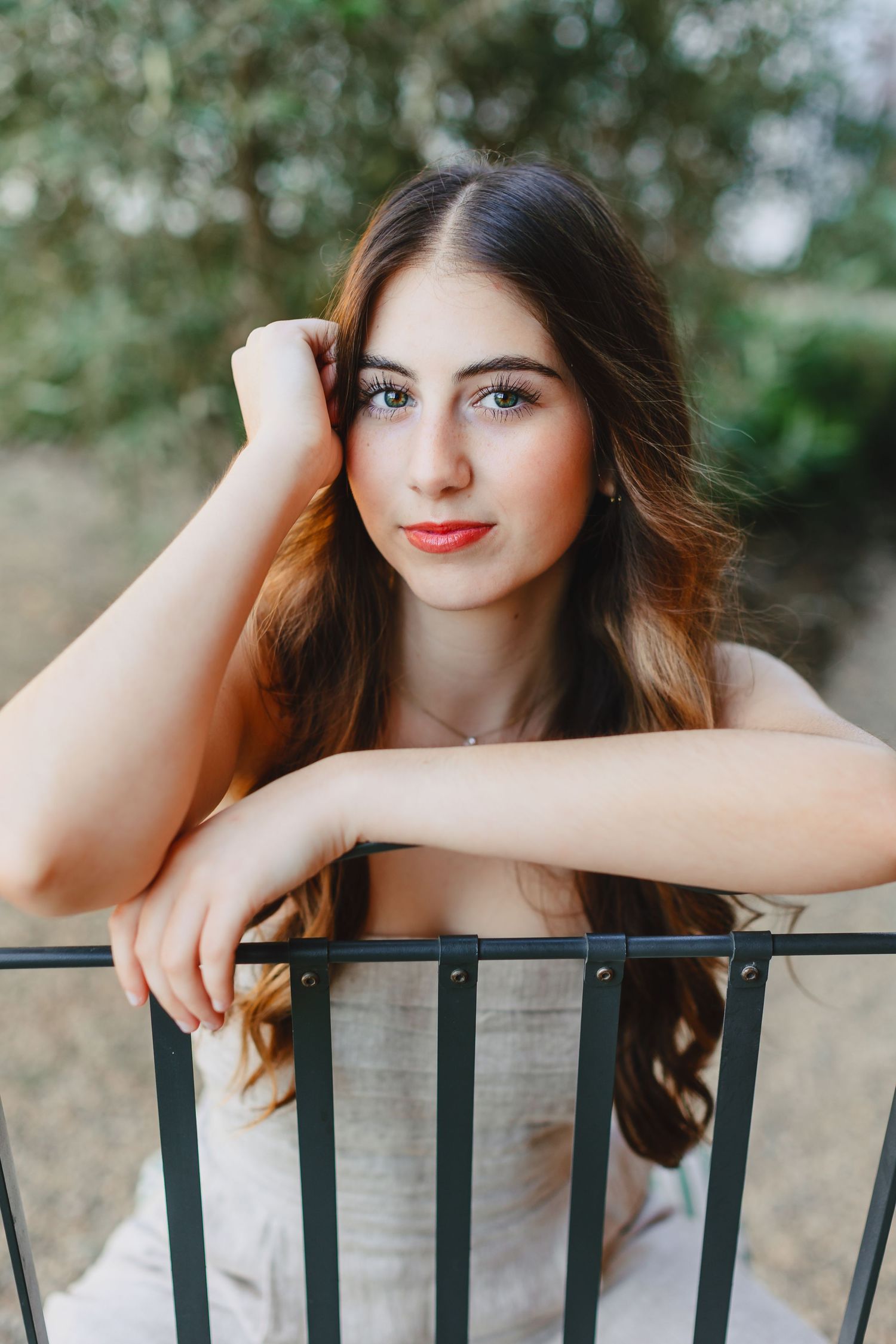 Young brunette woman leaning on metal chair back outdoors with soft green foliage background.