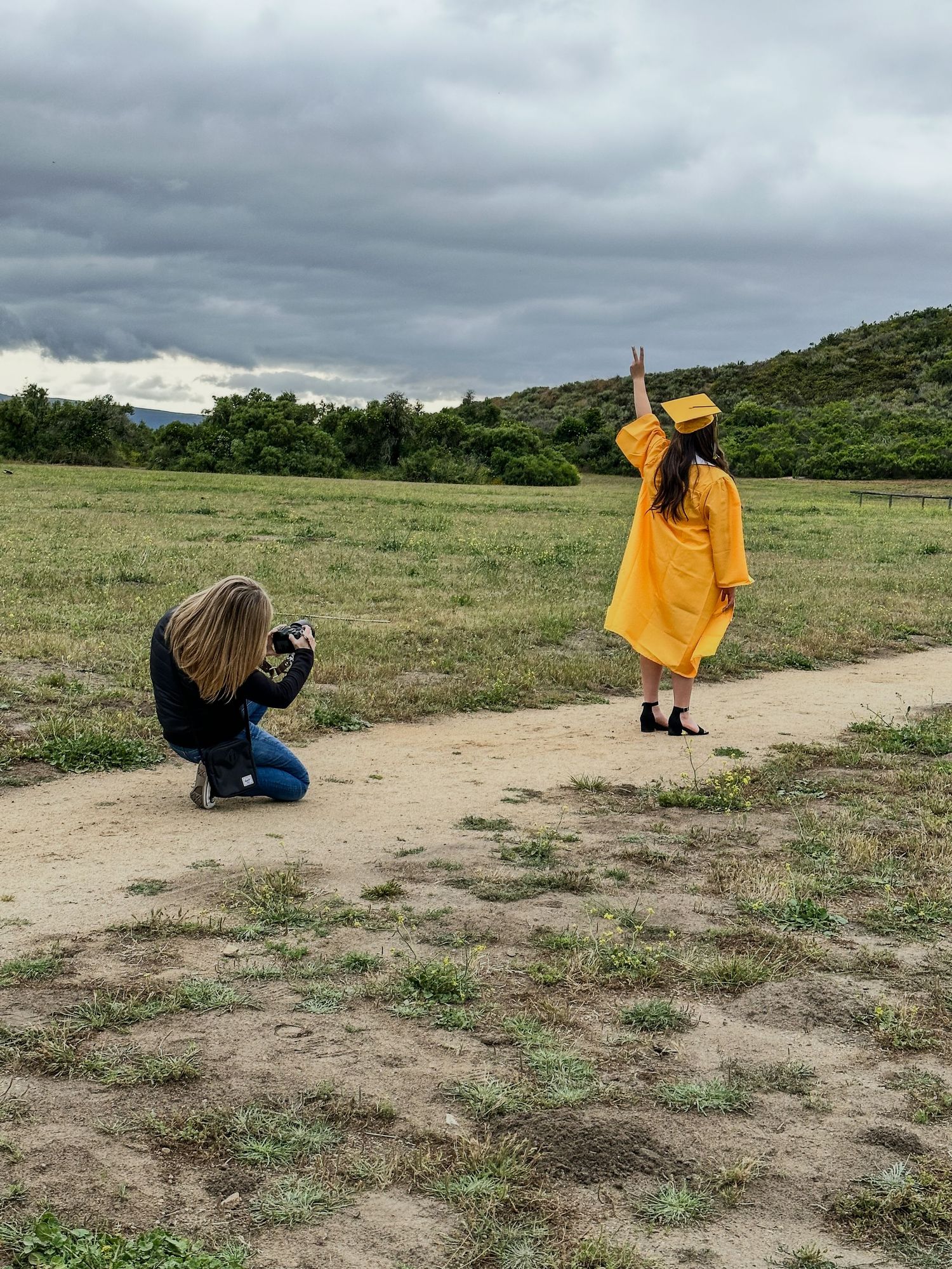 Blonde photographer kneels to photograph graduate in yellow cap and gown in open field.