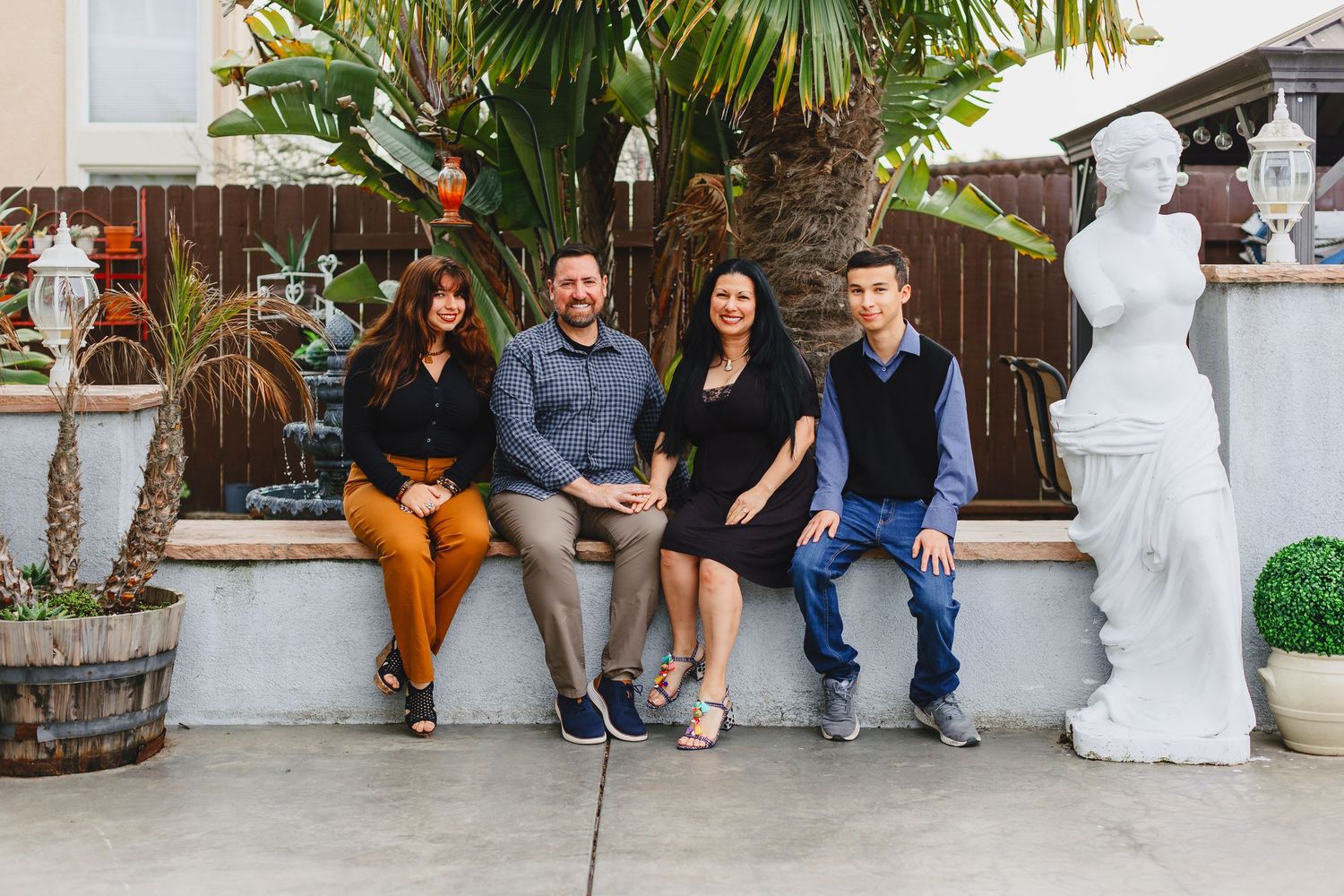 A group of young friends sitting outdoors on a patio bench next to a decorative white statue and potted plants.