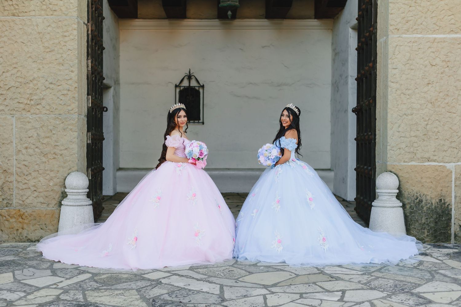 Two elegant ball gowns in pink and white with full tulle skirts are displayed in an outdoor stone archway.