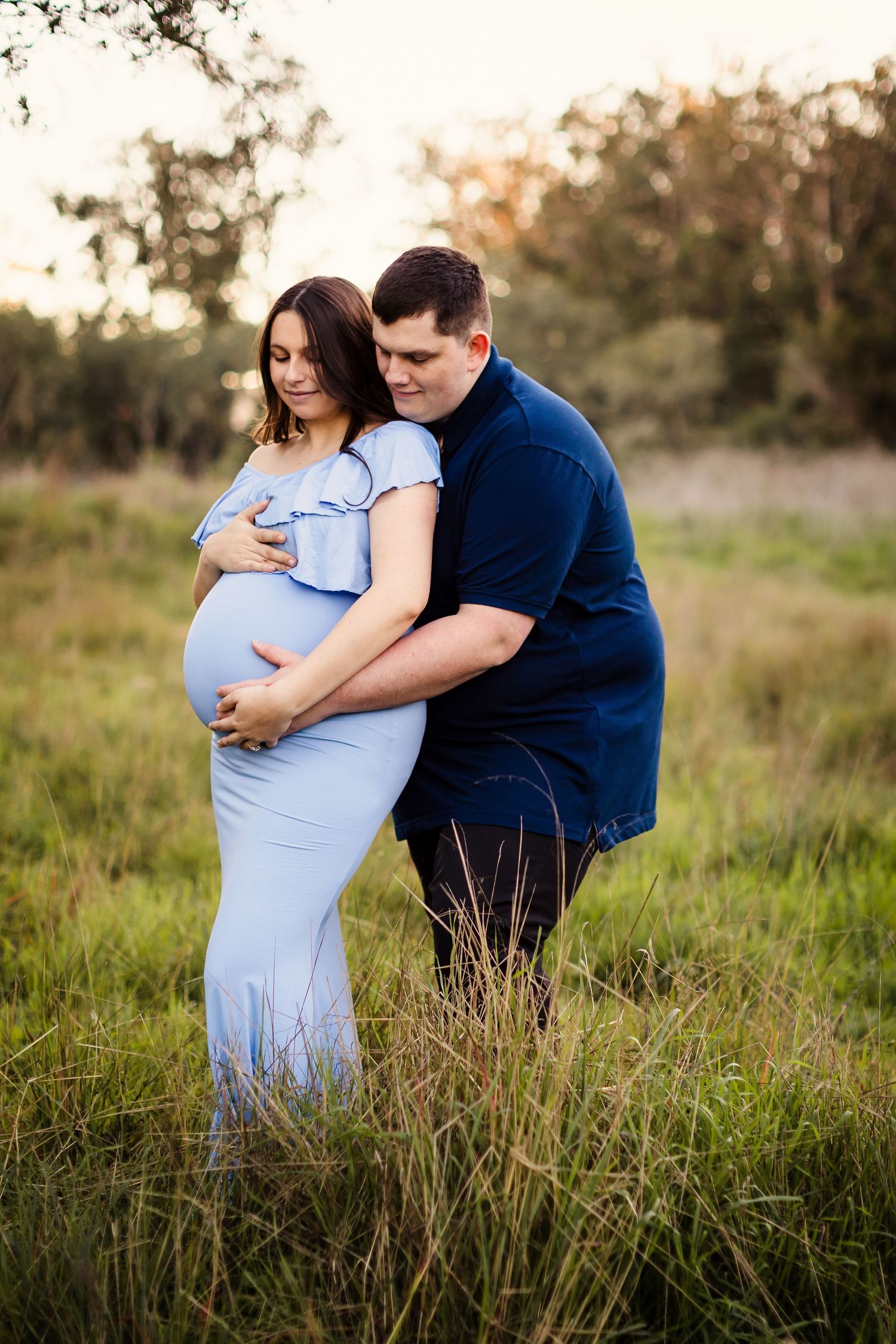 A couple embraces in tall grass during sunset, wearing a blue dress and navy shirt in an outdoor setting.