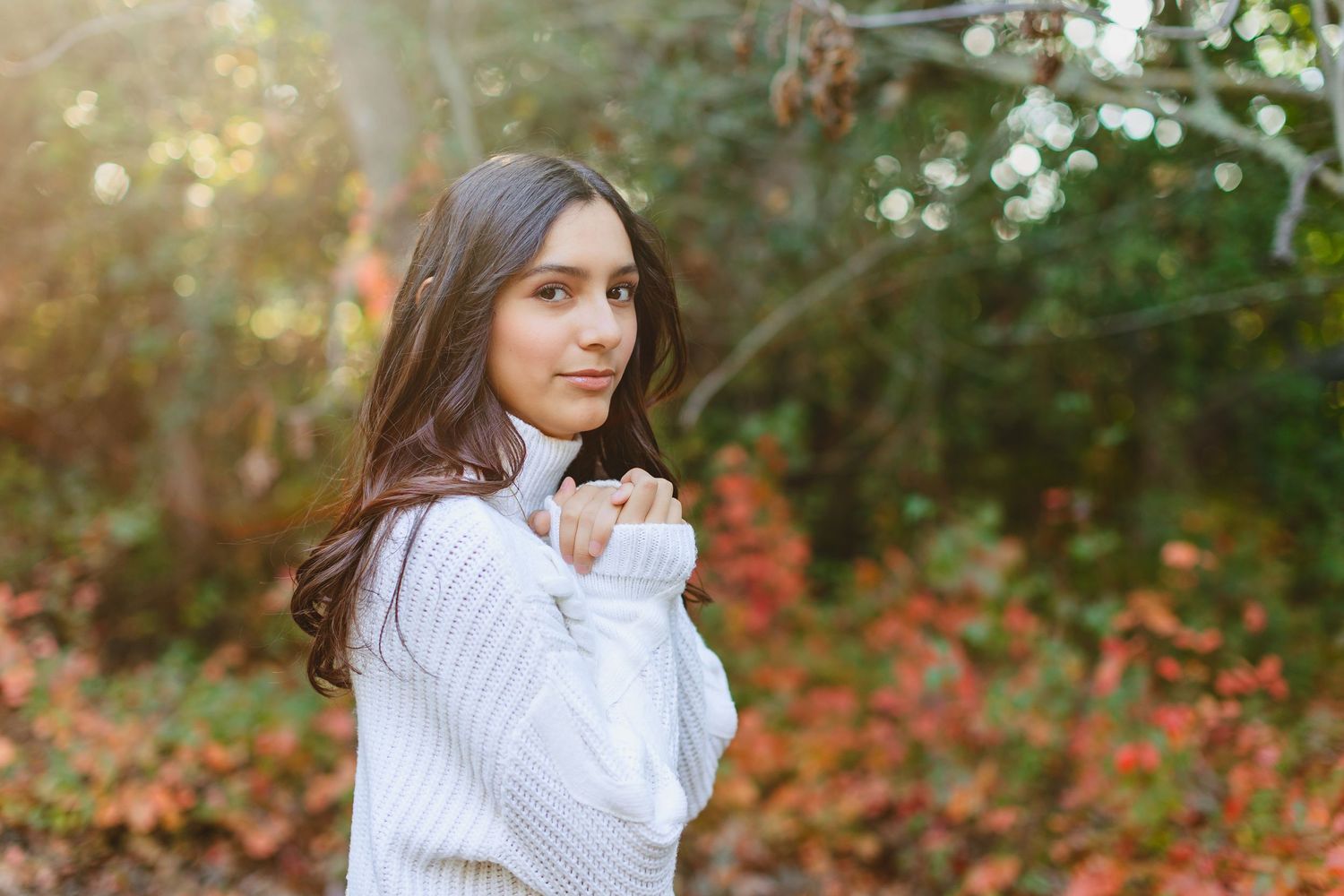 A person in a white sweater poses in an autumn forest setting with warm golden sunlight filtering through trees.