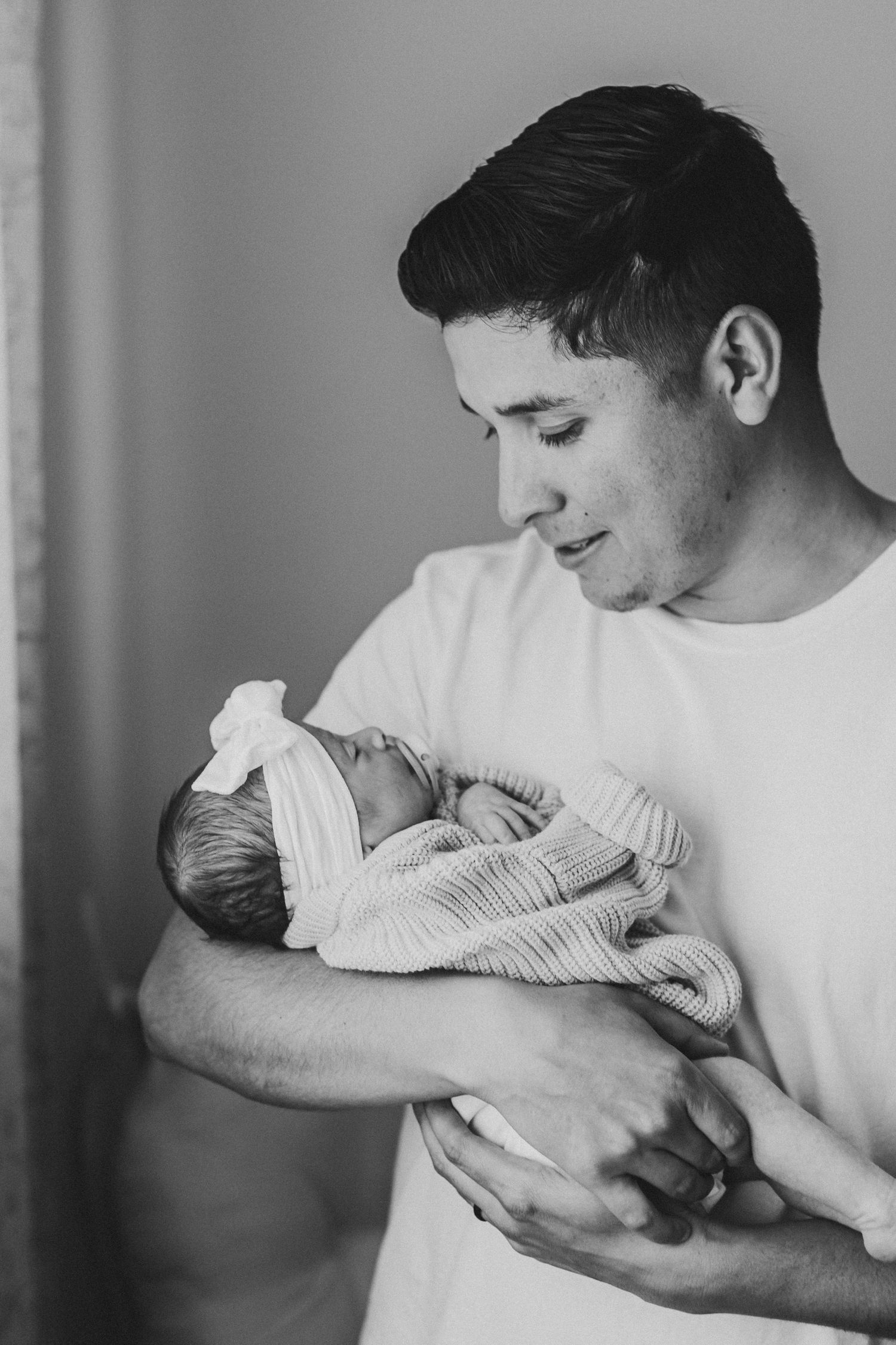 Black and white photo of a young man in a white t-shirt tenderly holding a sleeping newborn baby.
