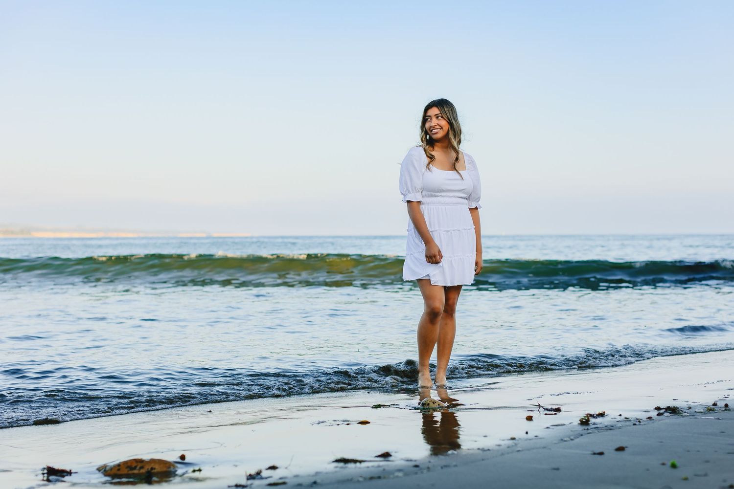 Young woman in white dress standing on a beach shoreline with ocean waves in the background.