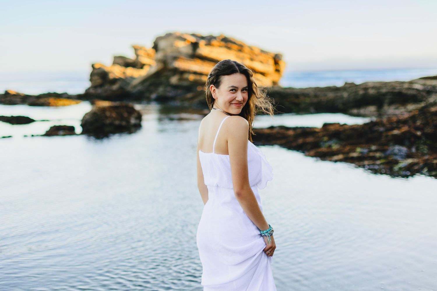 Person in white dress poses by rocky coastline during golden hour.