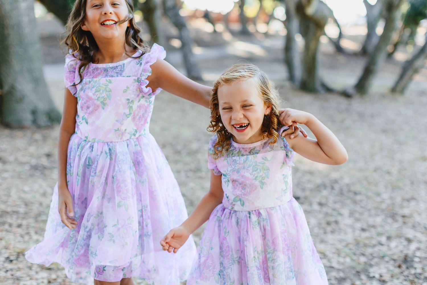 Two children in matching purple and white floral dresses laugh while playing outdoors.
