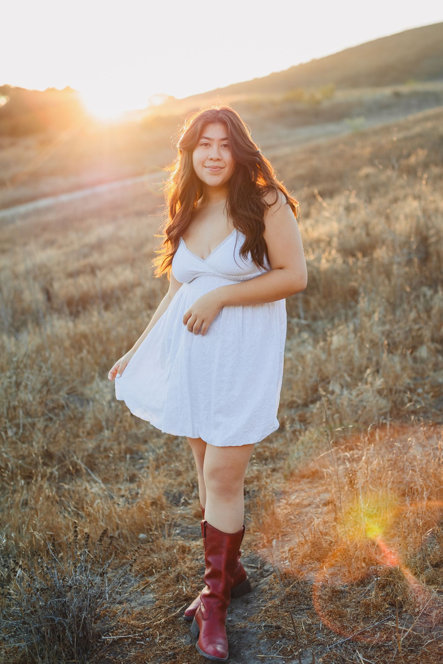 Woman in white sundress and red boots poses in golden field at sunset.