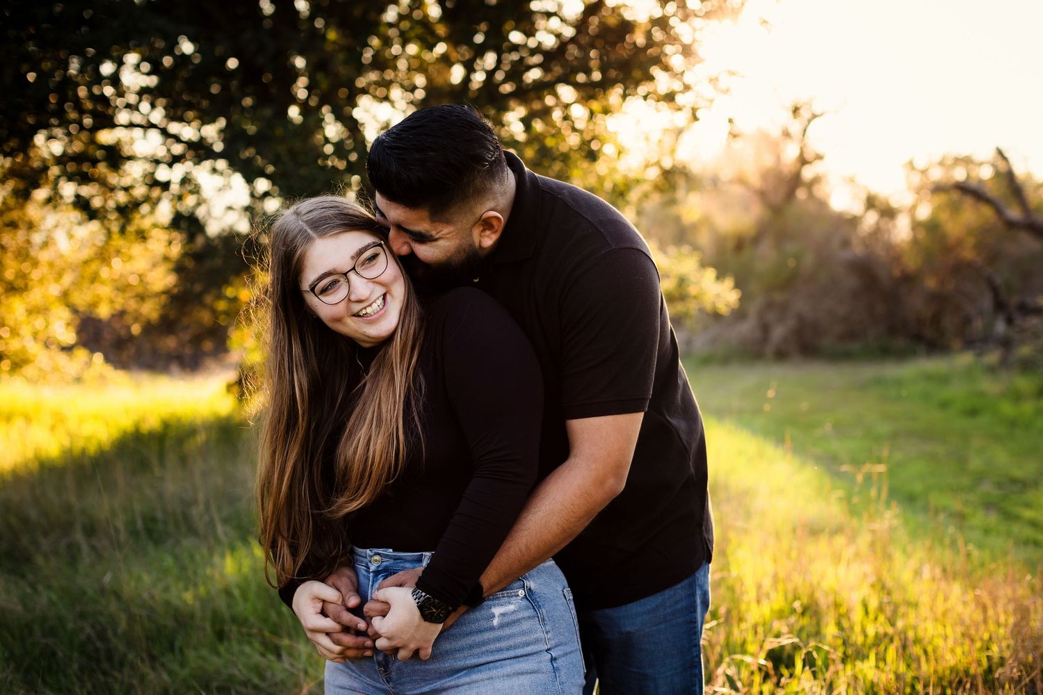 A couple embraces in a sunlit field during golden hour while looking at a phone together.