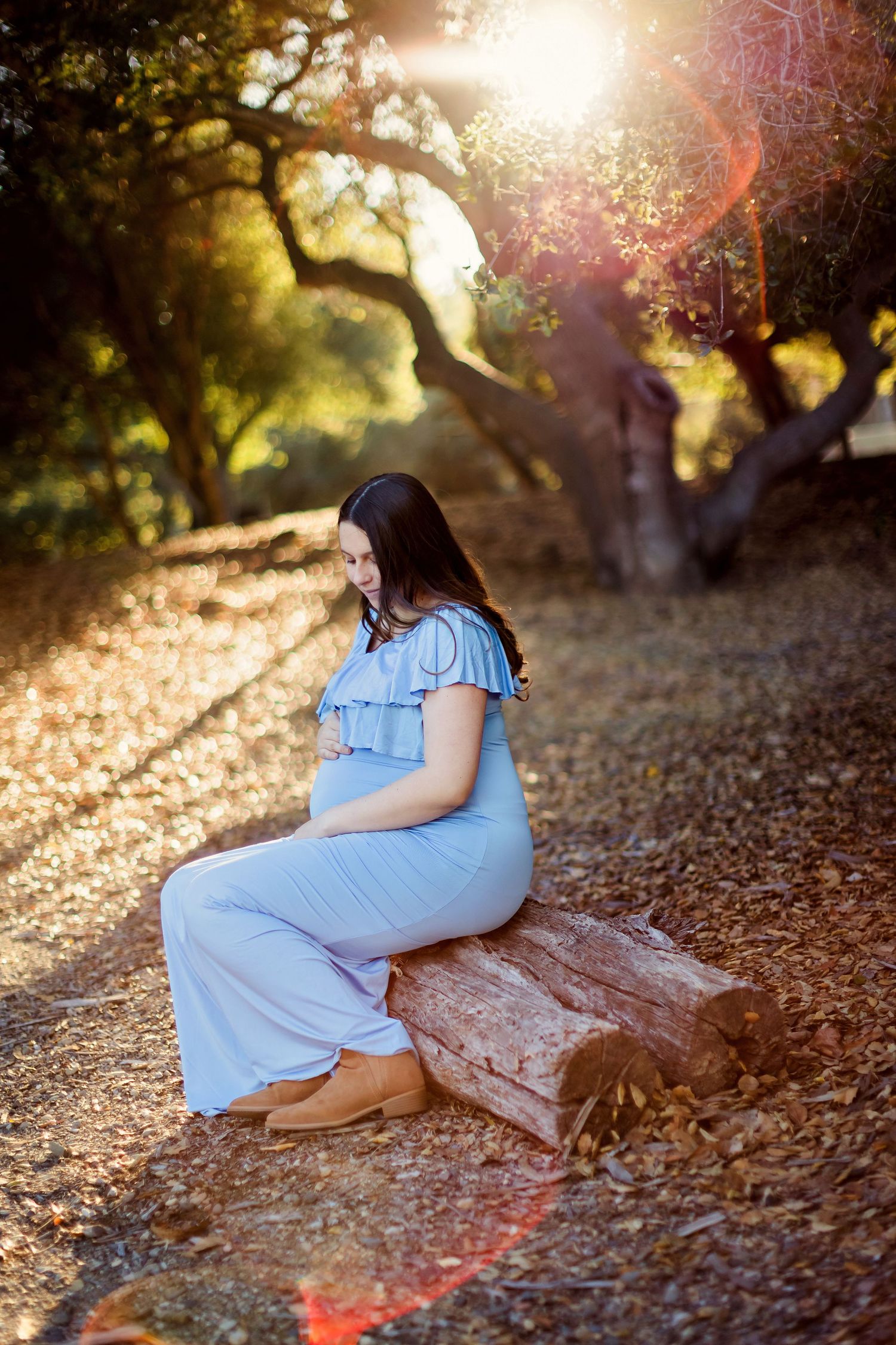 A woman in a light blue dress sits on fallen autumn leaves during golden hour sunset lighting.