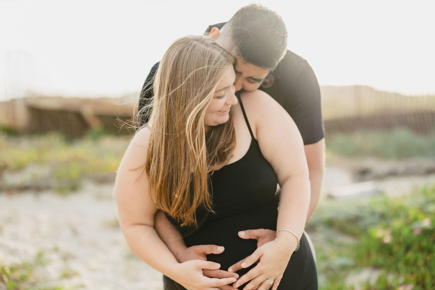 Couple embracing outdoors during maternity photo shoot, woman in black dress cradling baby bump at golden hour.
