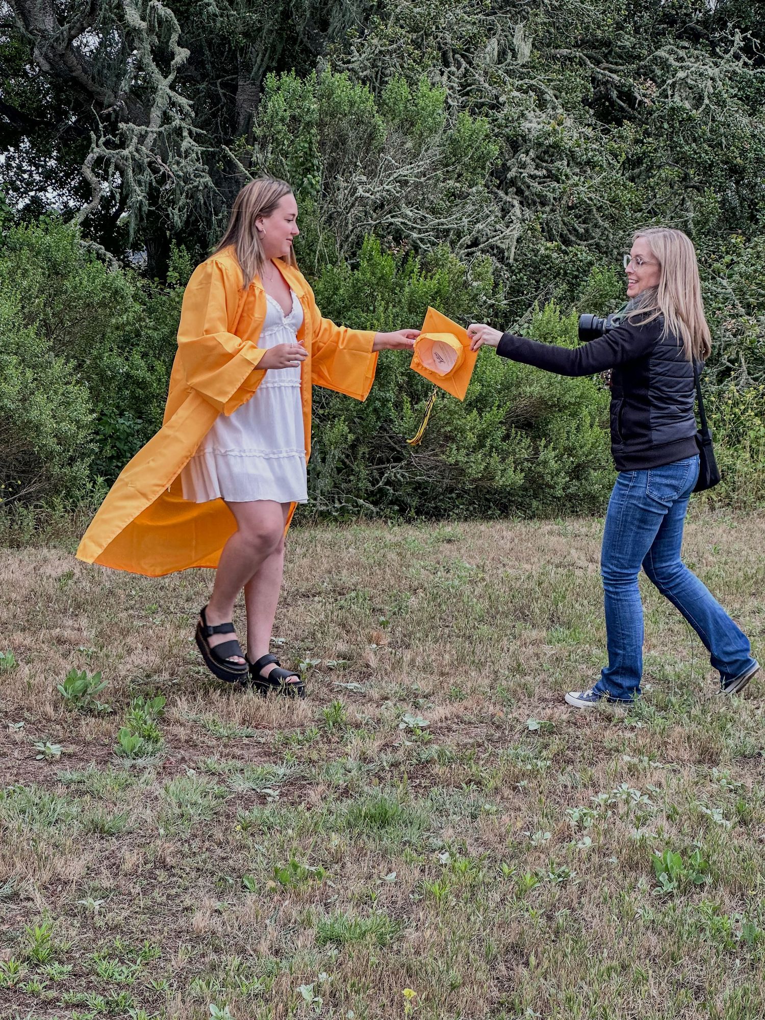 Two women outdoors exchanging an orange fabric accessory, one wearing a yellow kimono over a white dress.