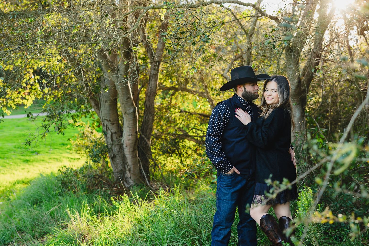 Couple embracing romantically under lush green trees in a sunlit outdoor setting, man wearing black cowboy hat and woman in dark outfit.